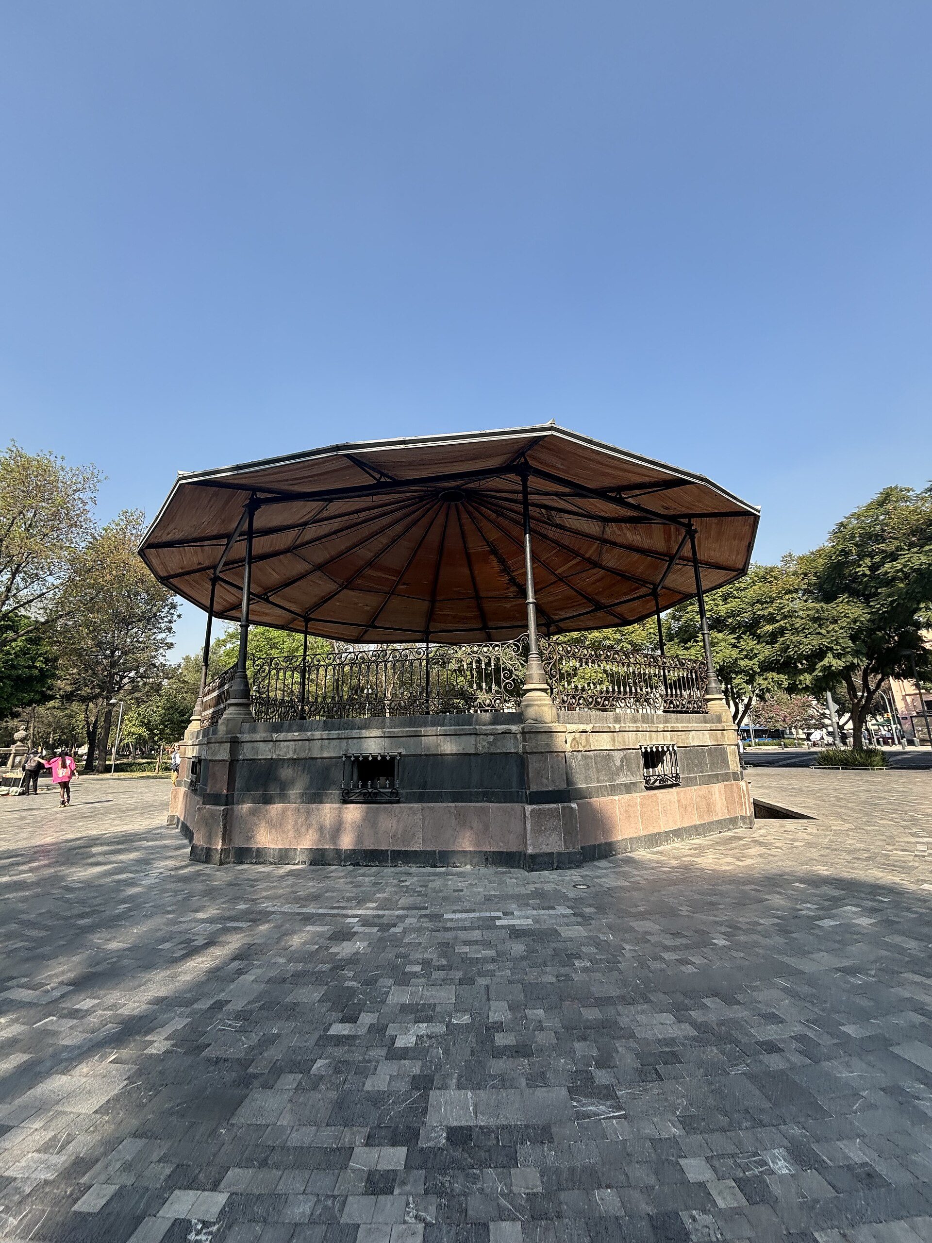 An outdoor gazebo with a large umbrella, surrounded by trees and a clear blue sky.