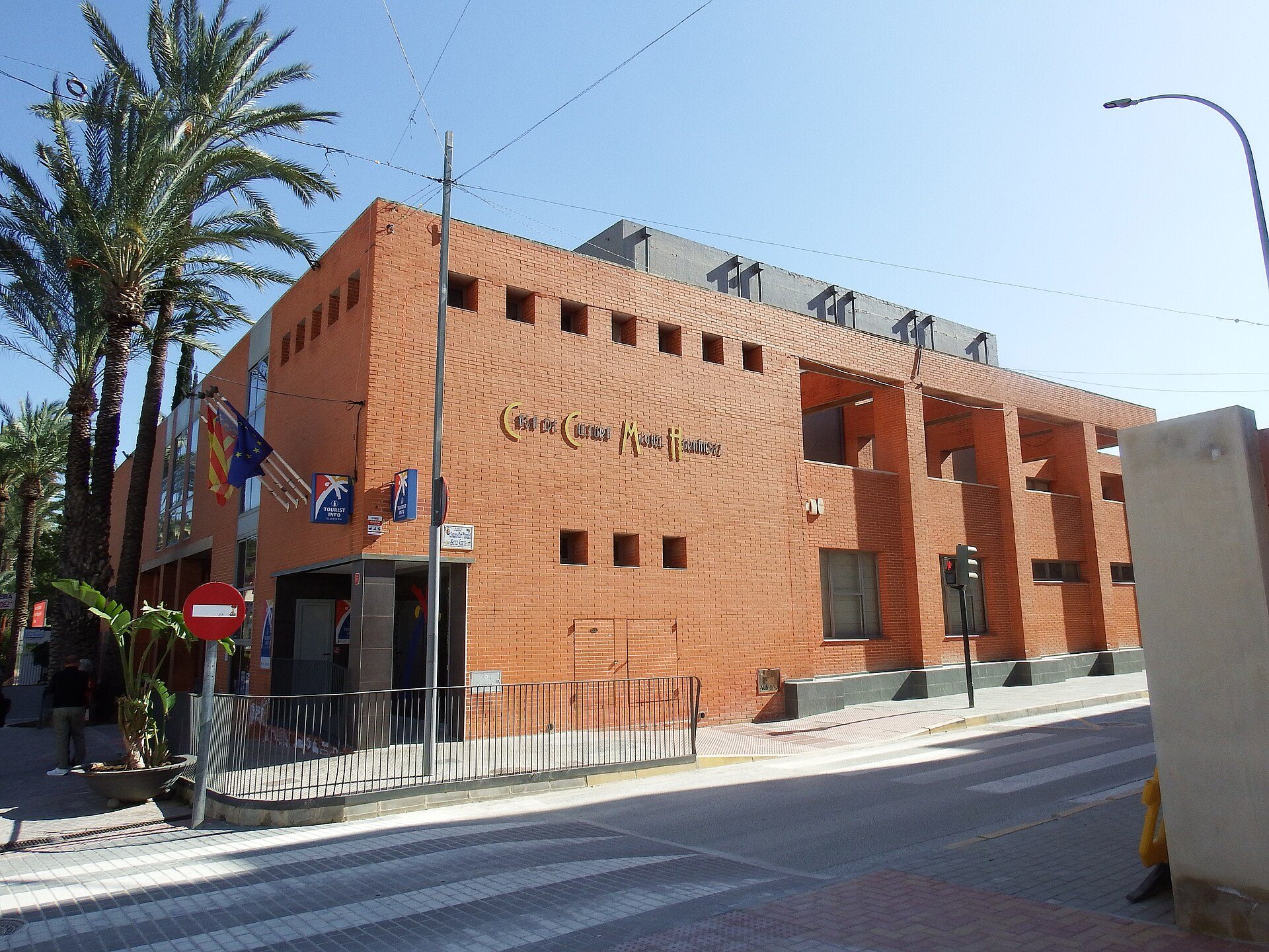 Red brick building with palm trees, modern design, large windows, and a rooftop terrace.