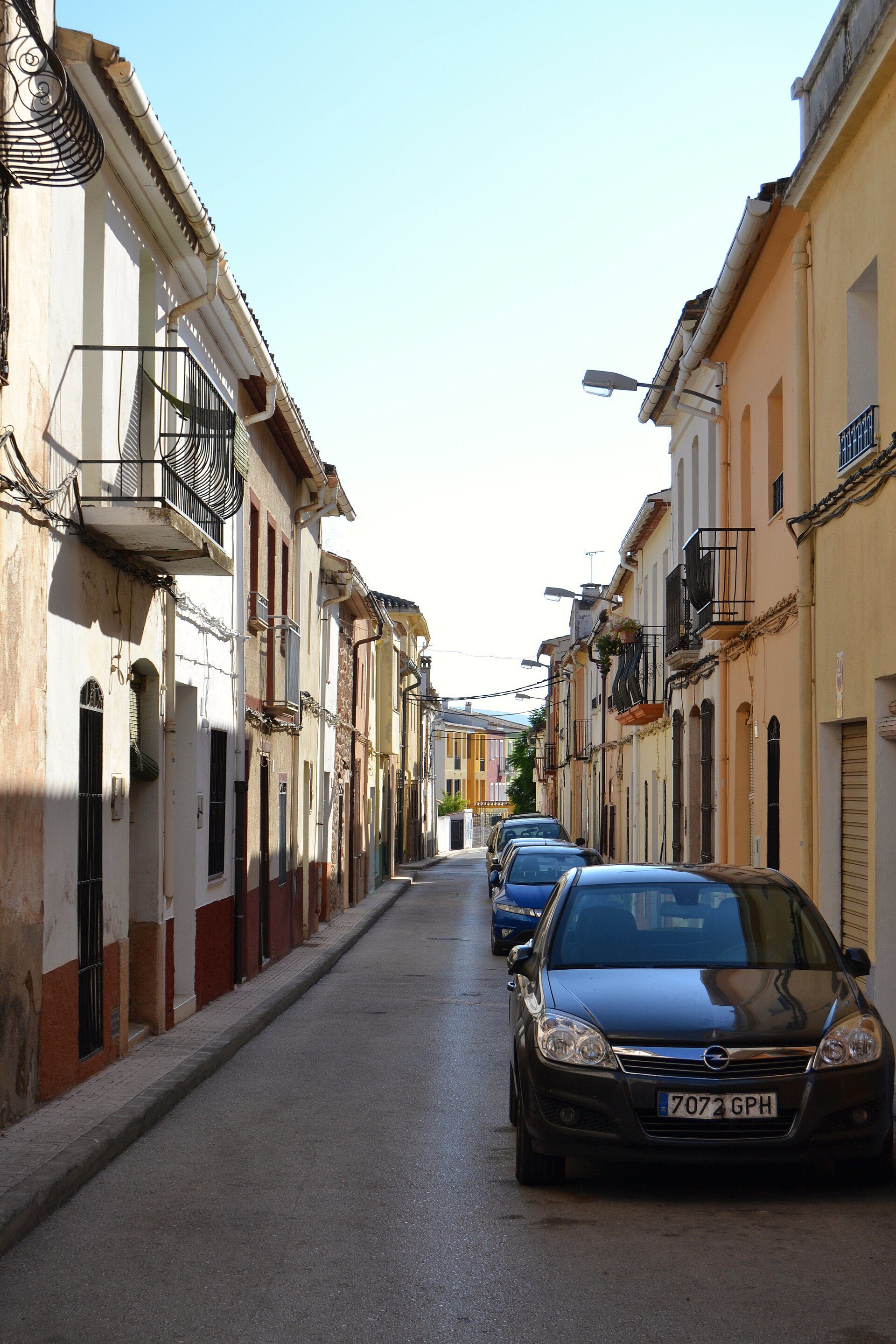 Narrow street with parked cars, colorful buildings, clear blue sky.