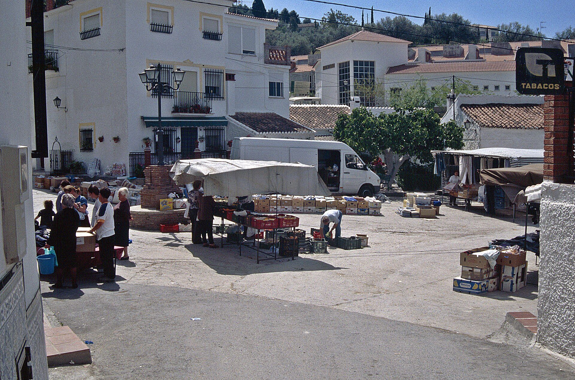A bustling outdoor market with vendors, boxes, and a white van, set against a backdrop of white buildings and a clear sky.