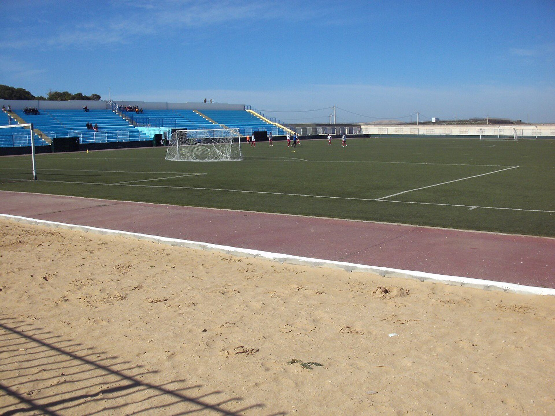 Alt text: Outdoor soccer field with blue seating, sandy area, and clear sky.