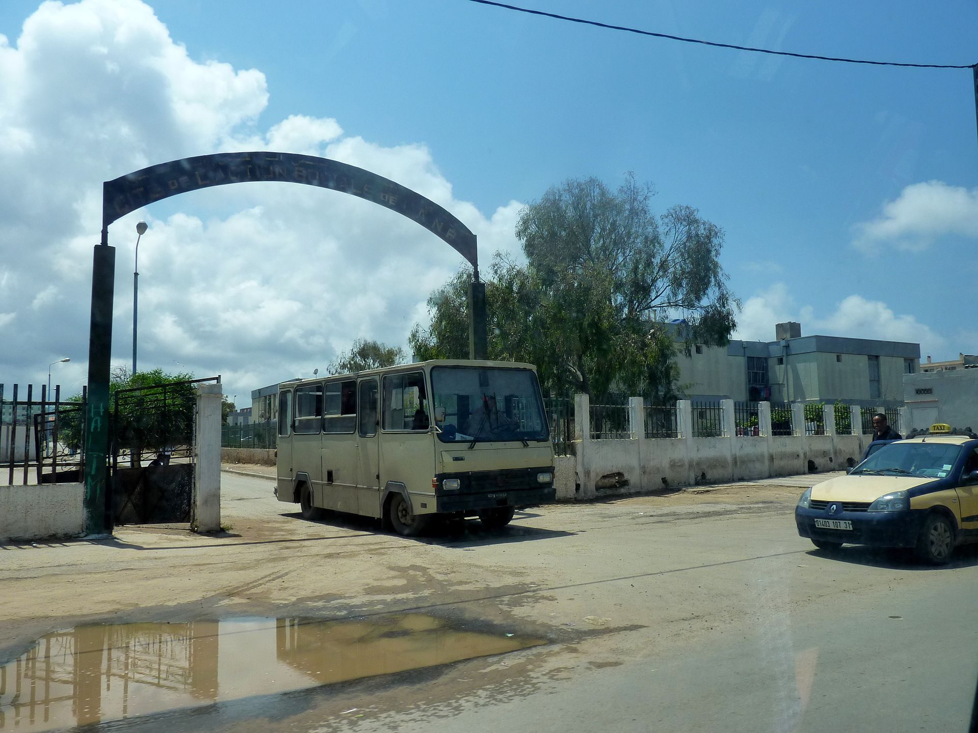A beige van is parked under an archway with a clear blue sky and trees in the background.