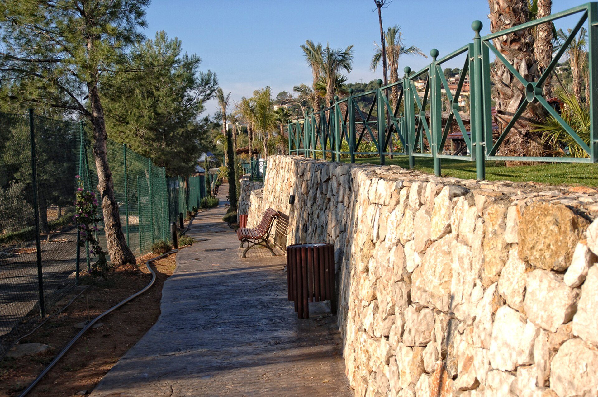 Alt text: Stone pathway with benches, green fence, and trees, leading to a scenic view.