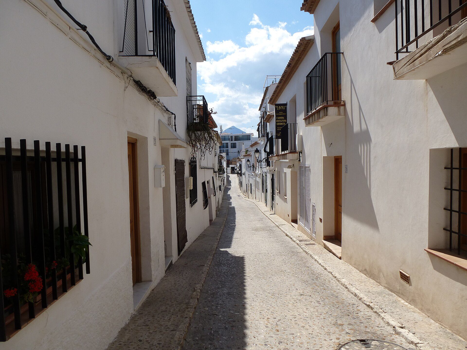 Narrow alley with white buildings, balconies, and a clear blue sky.
