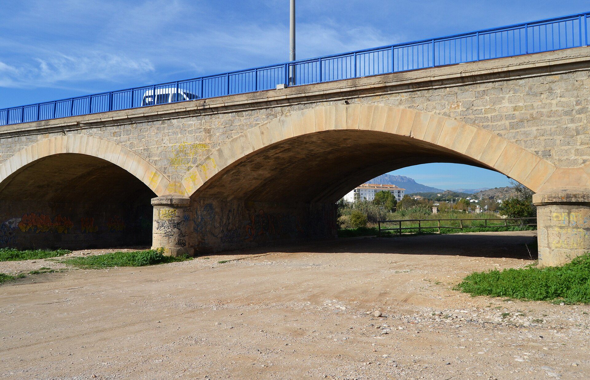 Alt text: Stone bridge with arched openings, blue railing, and a clear blue sky in the background.