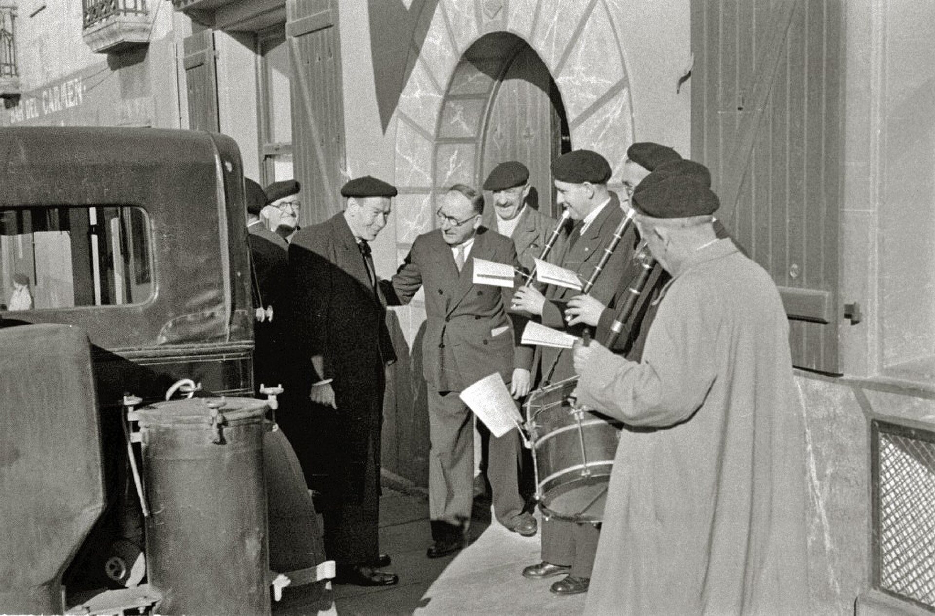 A black and white photo of a group of people outside a building, with a vintage car and musicians.