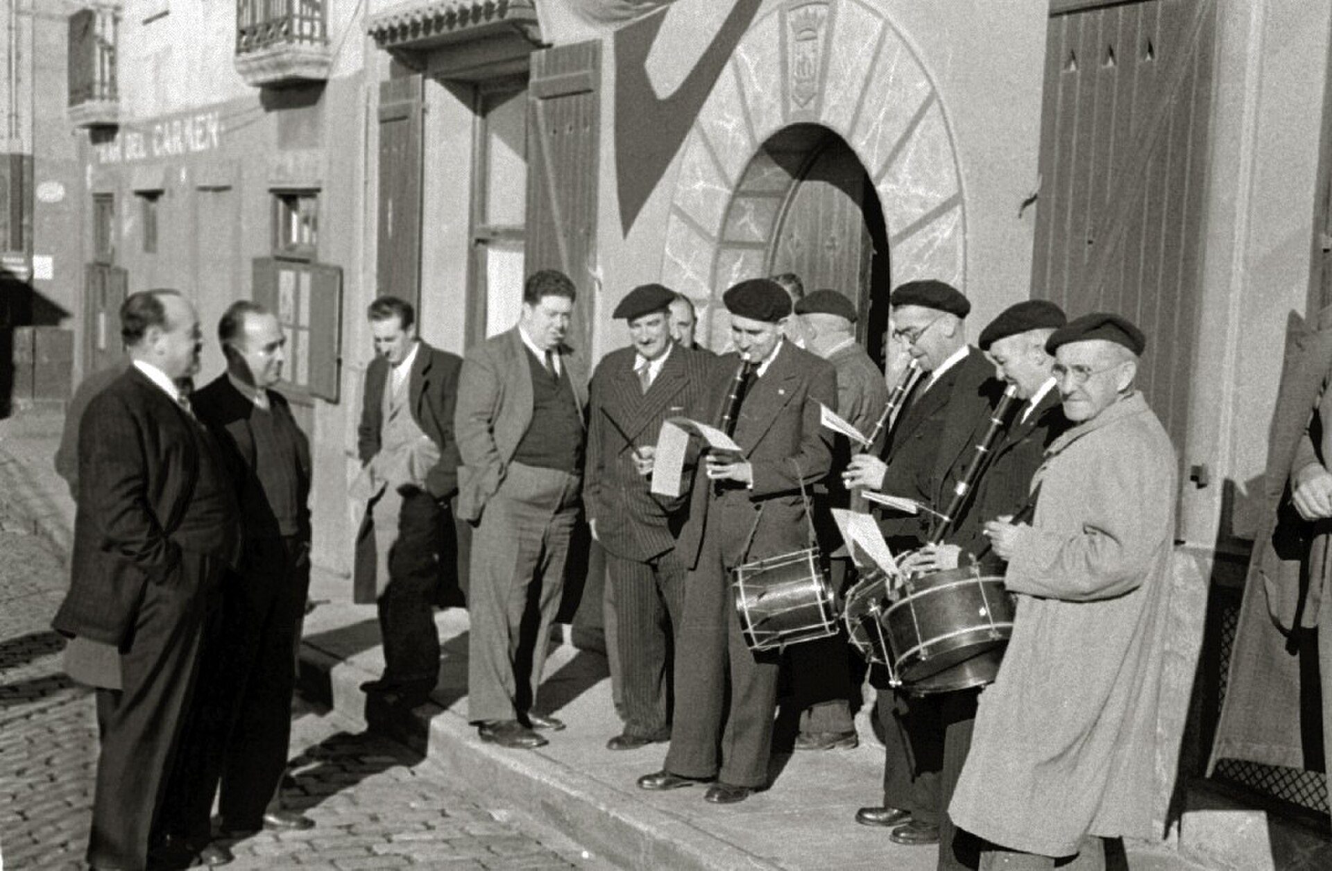 A black and white photo of a group of men in suits and musicians performing on a street.