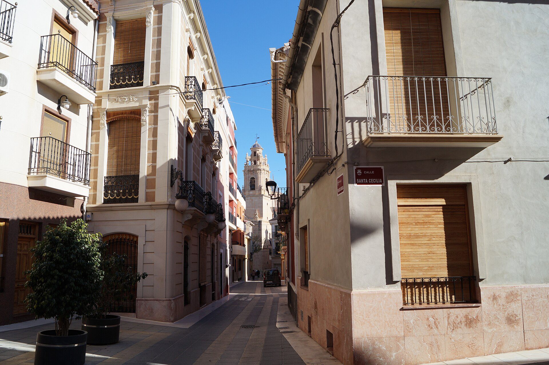 Narrow street with historic buildings, balconies, and a church tower in the distance.