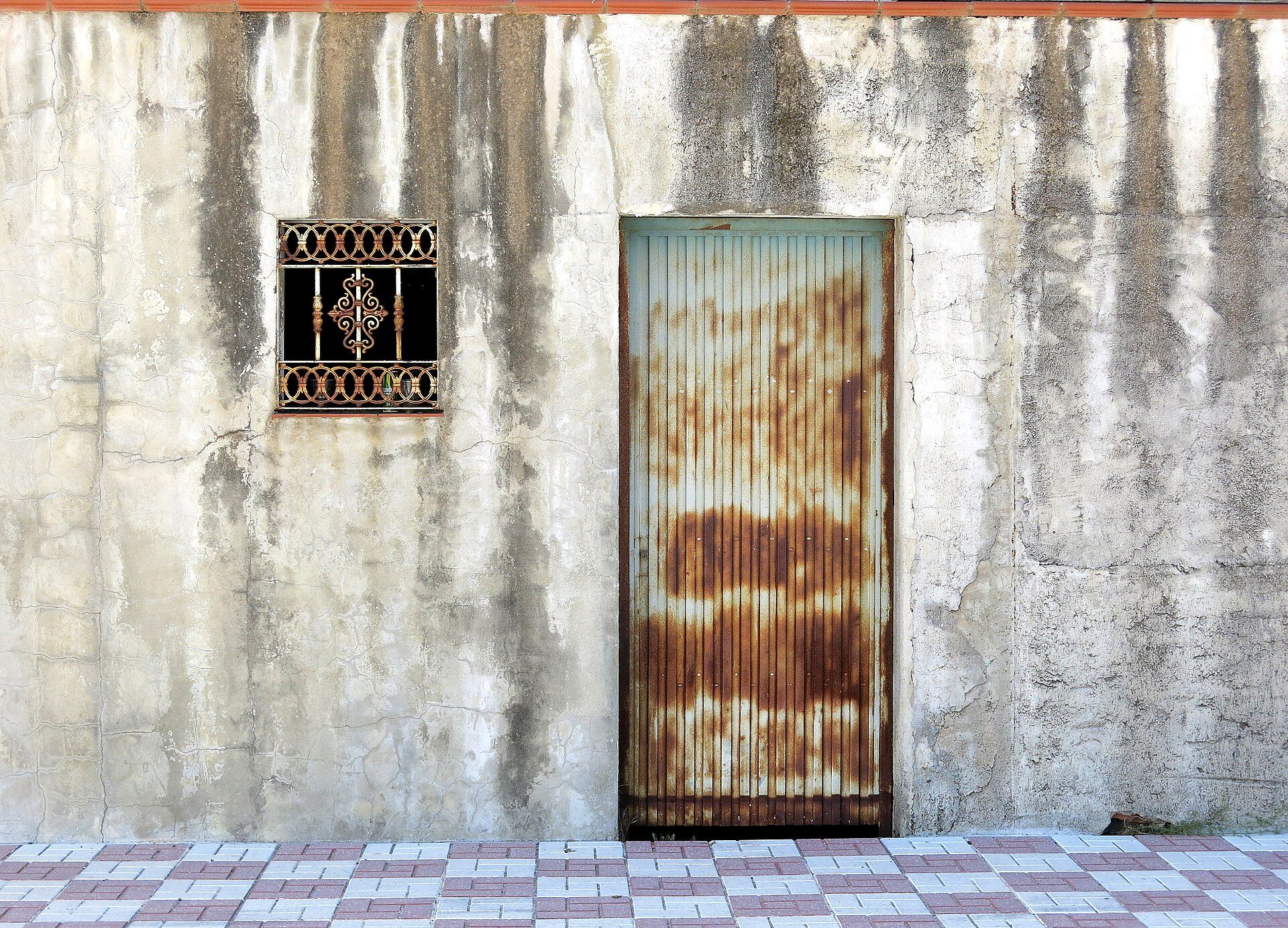 Old, weathered concrete wall with a rusty metal door and decorative window.
