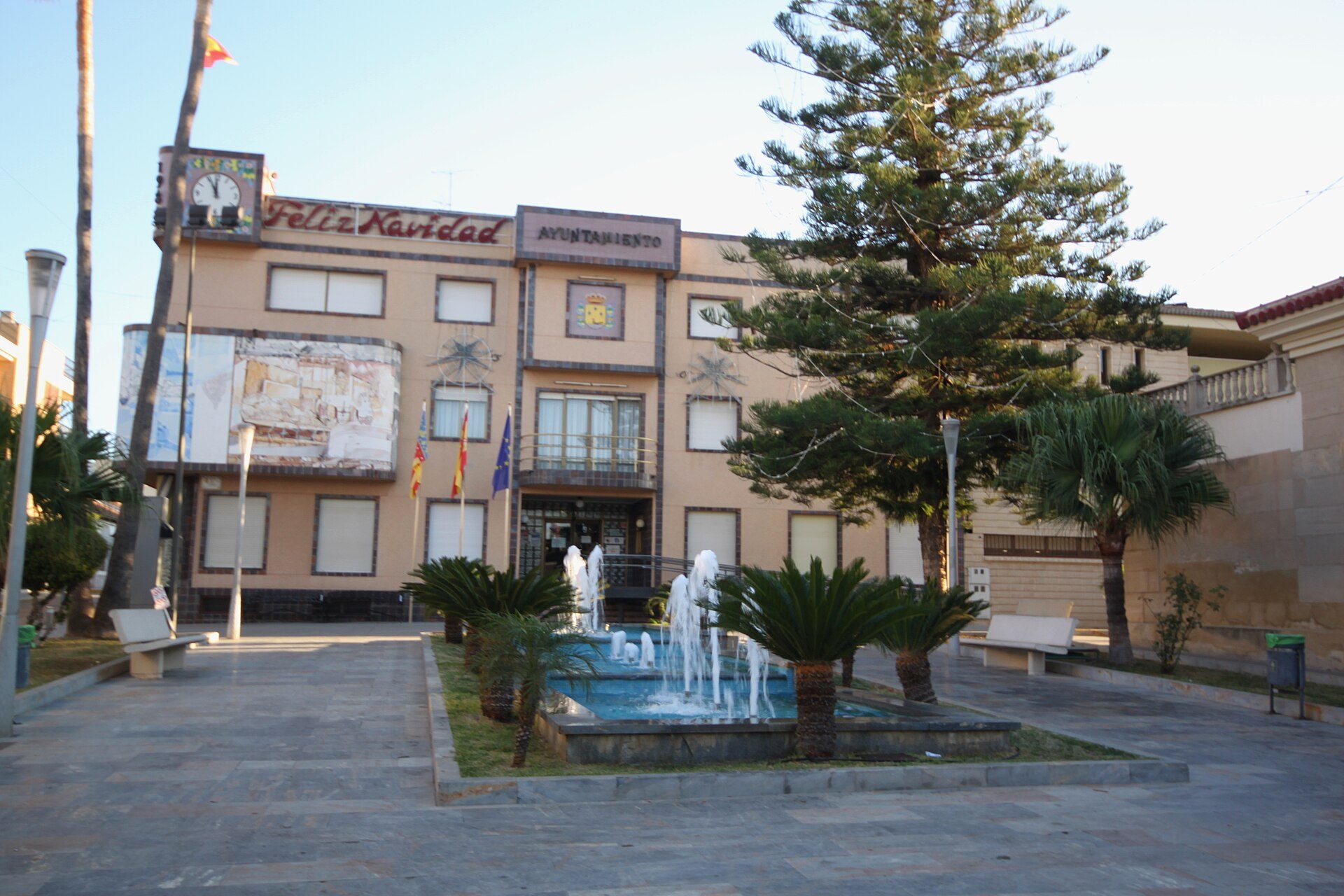 A courtyard with a fountain, surrounded by palm trees and a building with a clock.