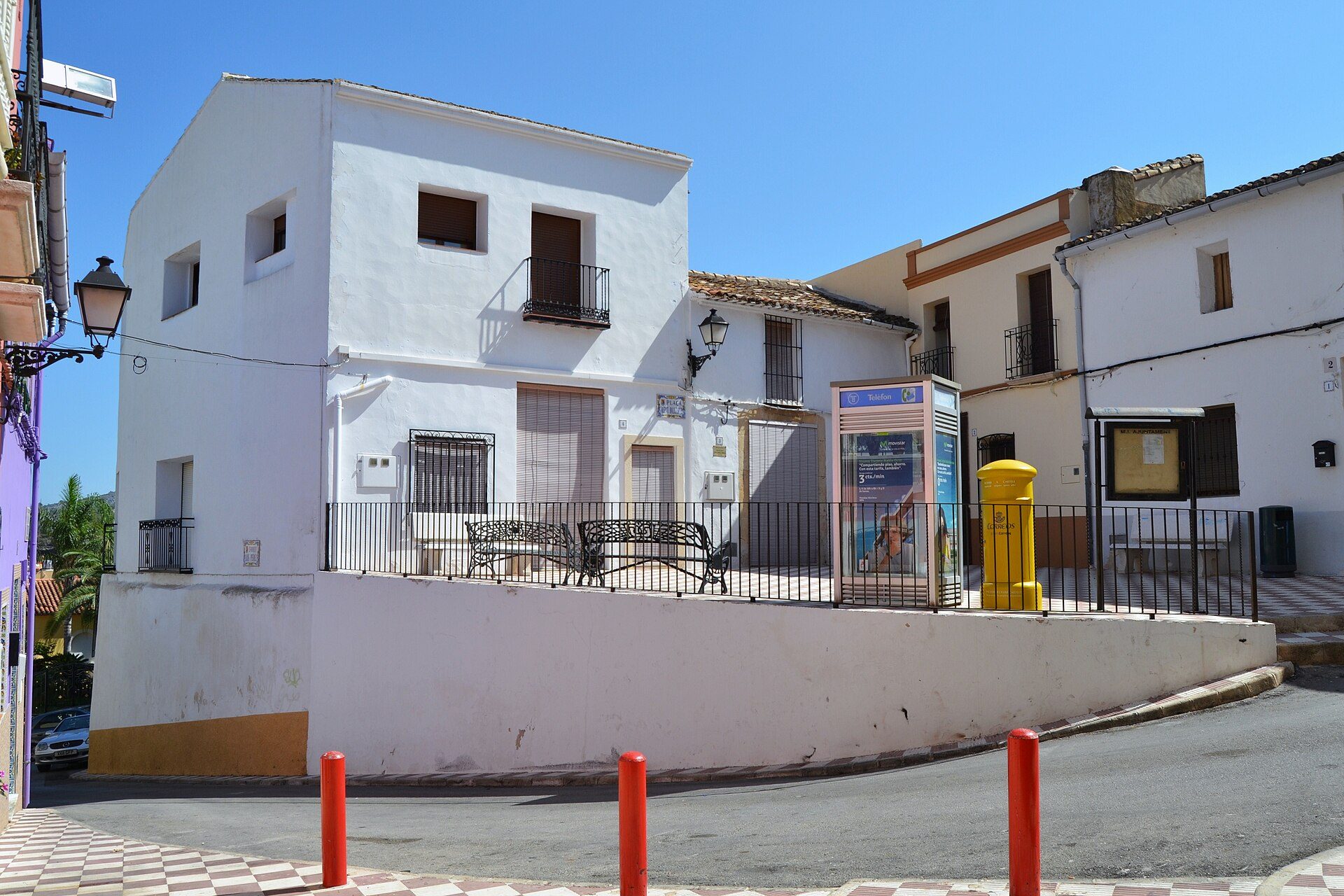 Two-story white building with balconies, street view, black metal railing, benches, and a yellow mailbox.