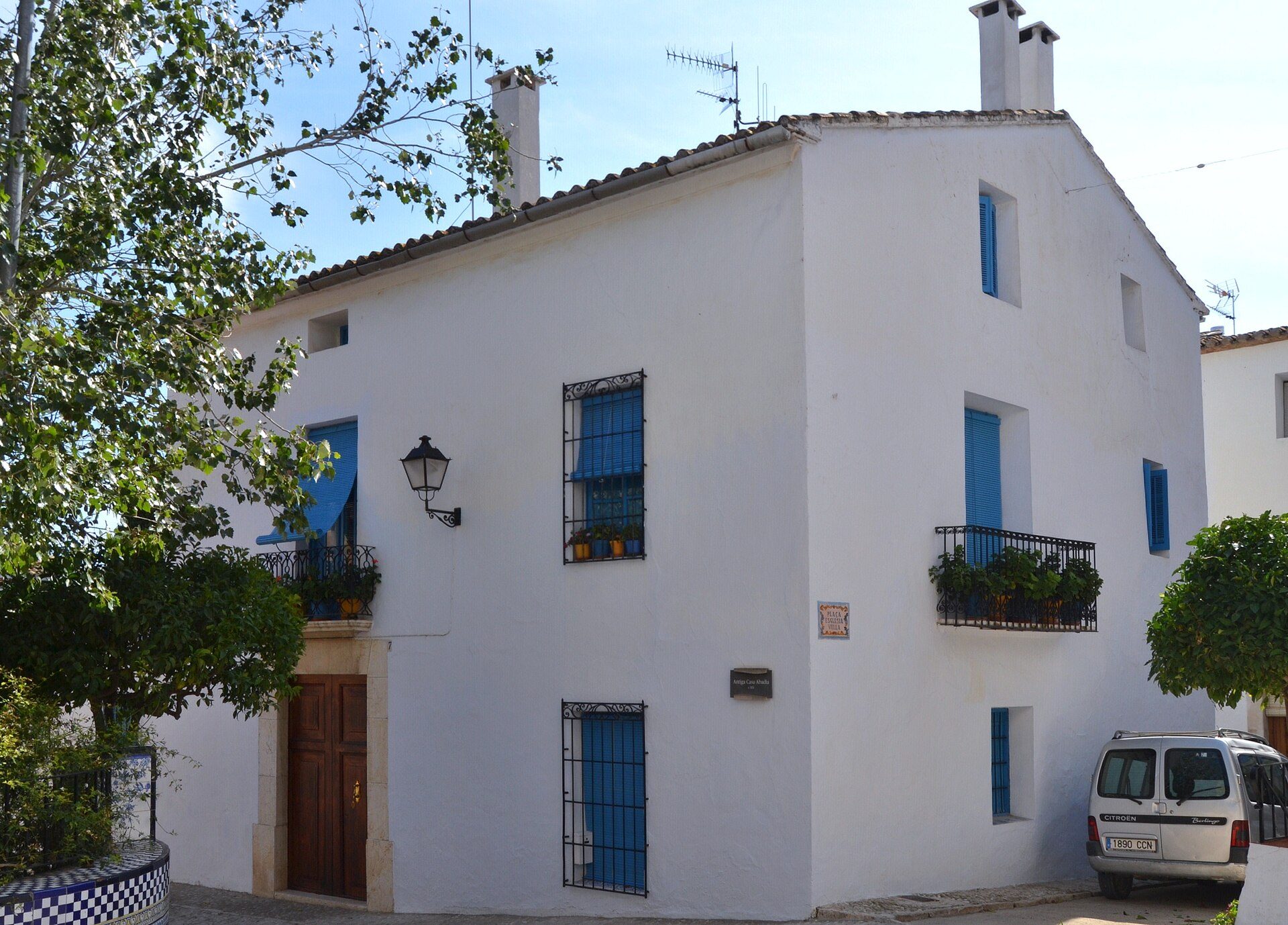 Two-story white house with blue shutters, balconies, and a small garden.