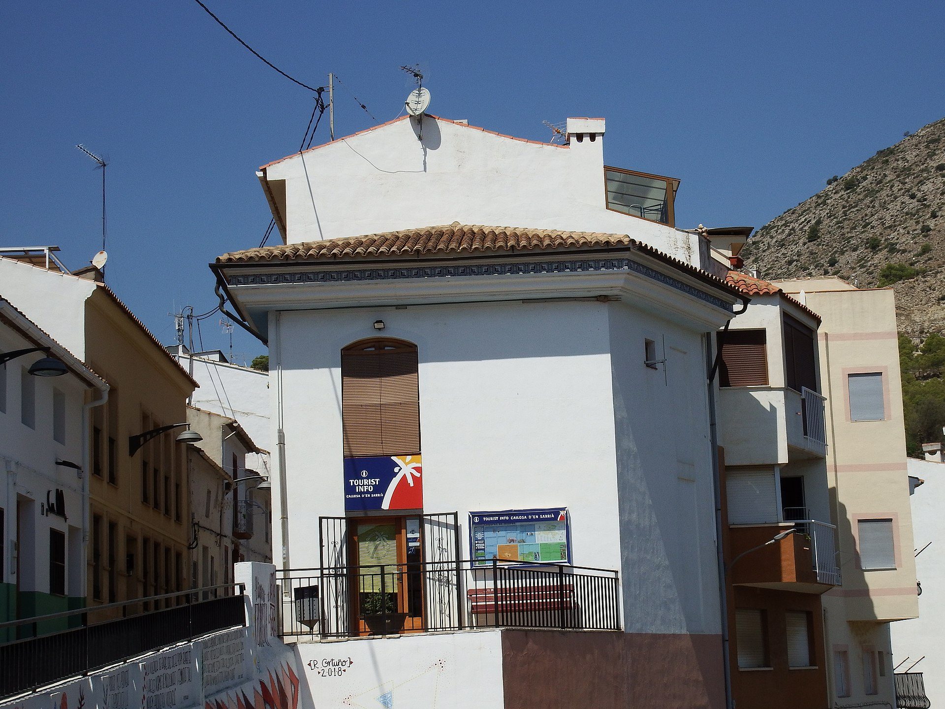 White building with a balcony, mountain view, satellite dishes, and a blue sign.