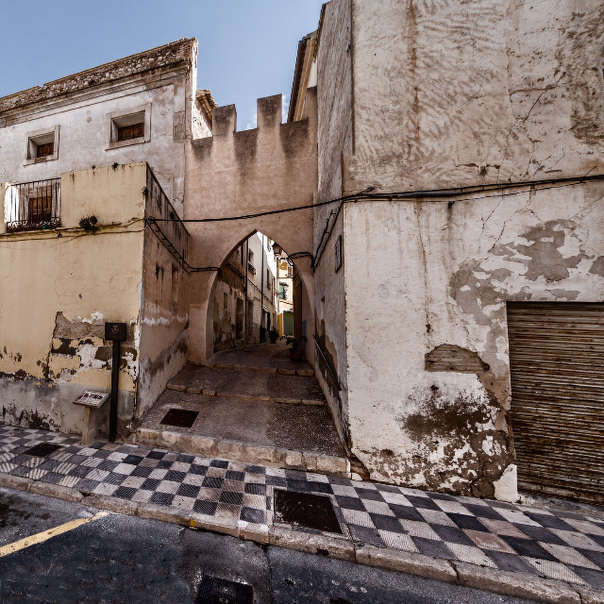 Narrow alley with archway, checkered pavement, old buildings, and a staircase.