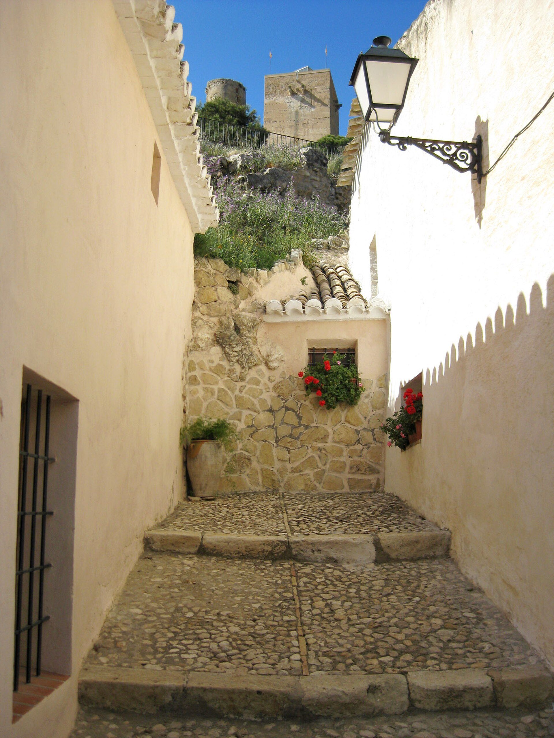 Cozy stone-paved alley with a view of a fortified hilltop, featuring a lantern and flower boxes.