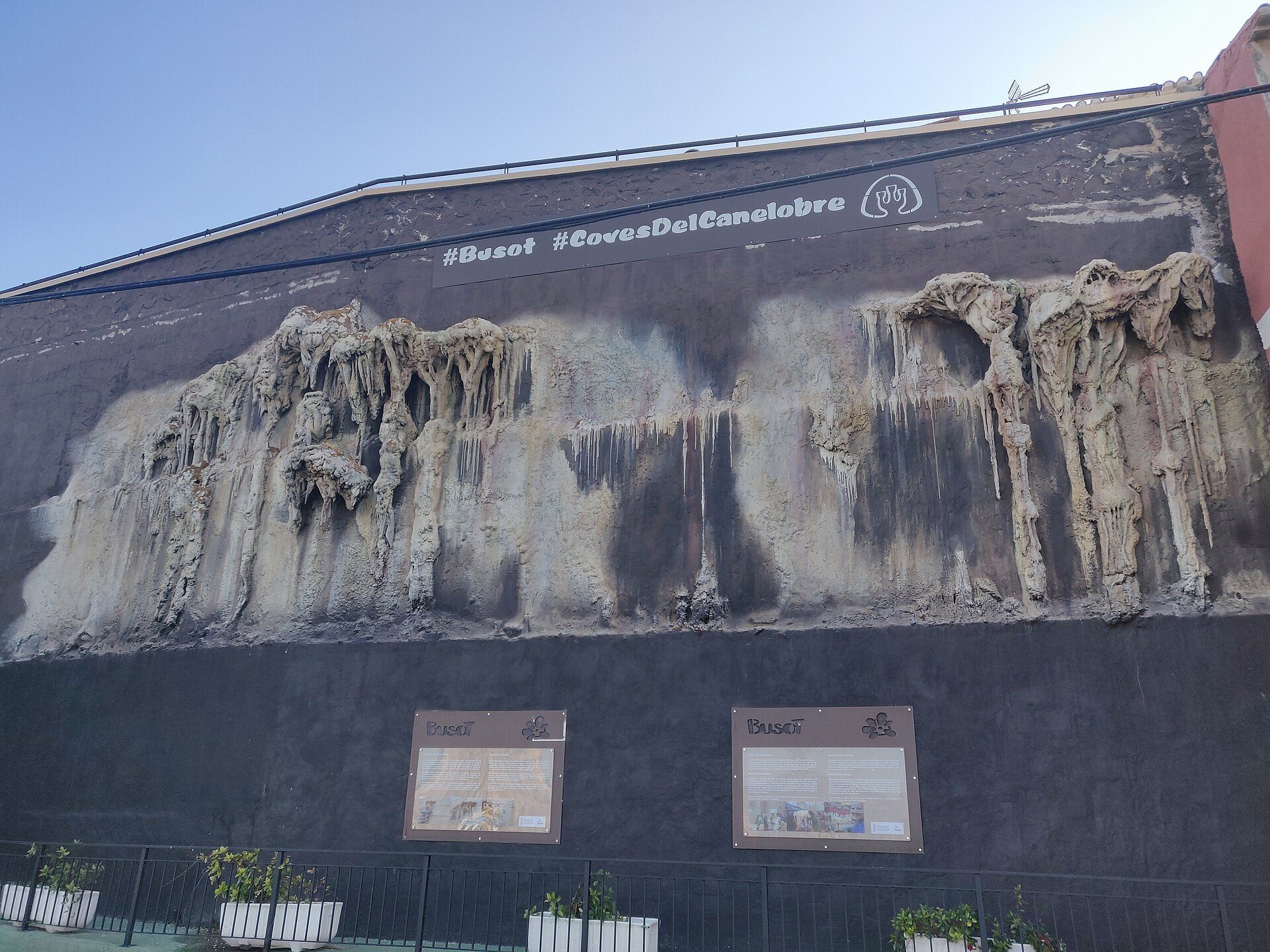 Alt text: Large mural on building wall with #Buzón and #CovesDelCanelobre hashtags, two information boards below.