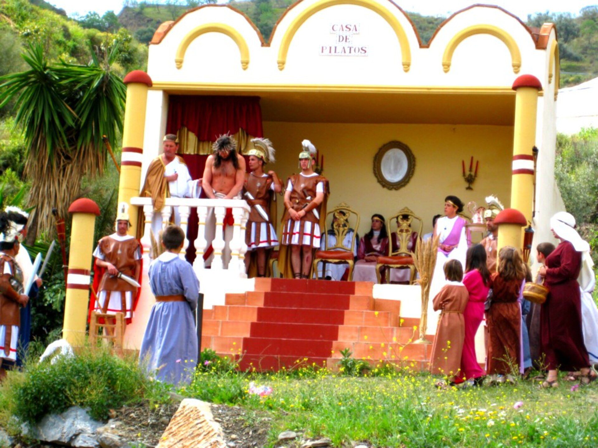 Alt text: Outdoor stage with Roman-themed performance, greenery, and audience.