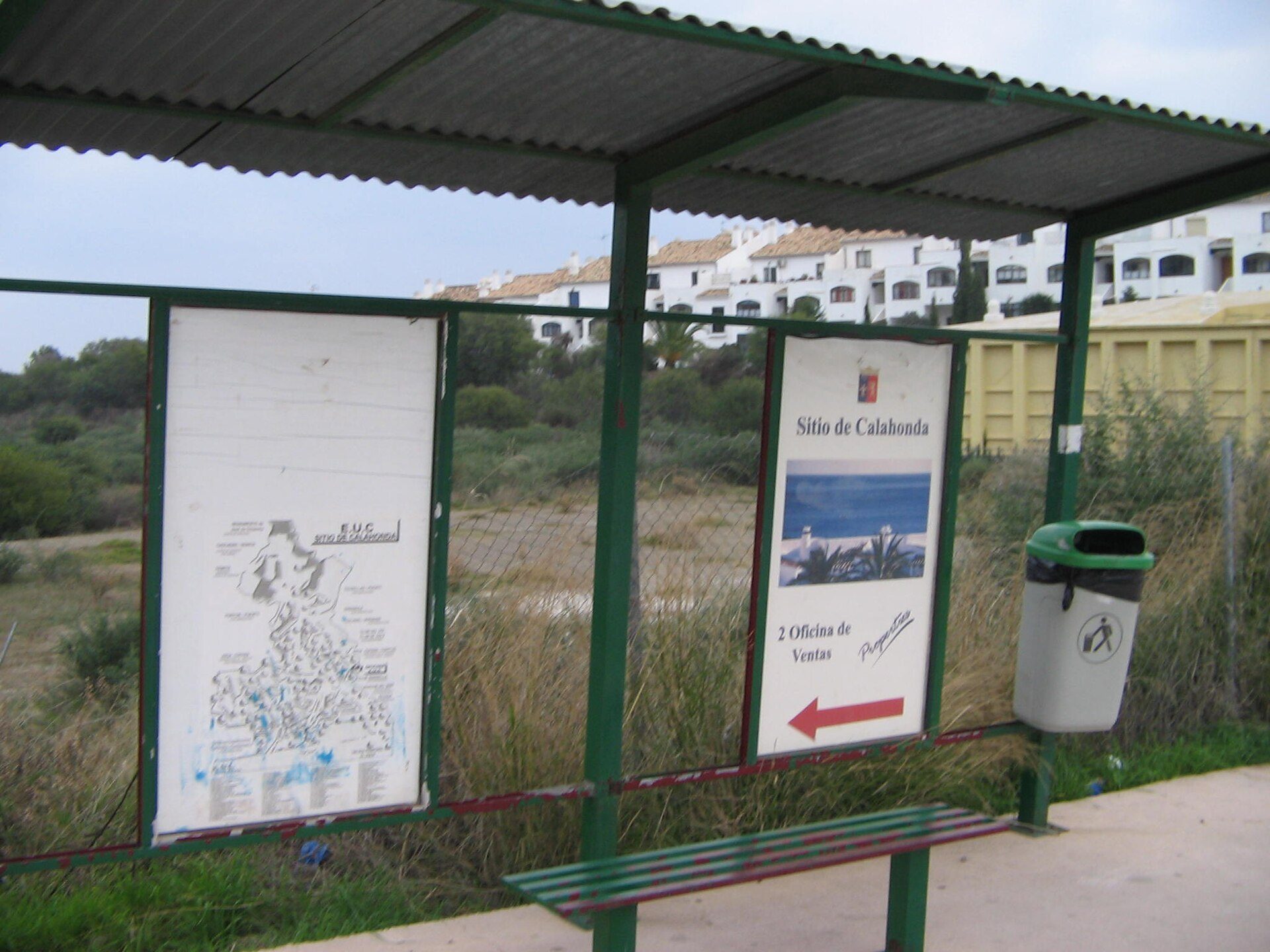 Alt text: Outdoor bus stop with map, bench, and trash bin, scenic view of buildings and greenery.