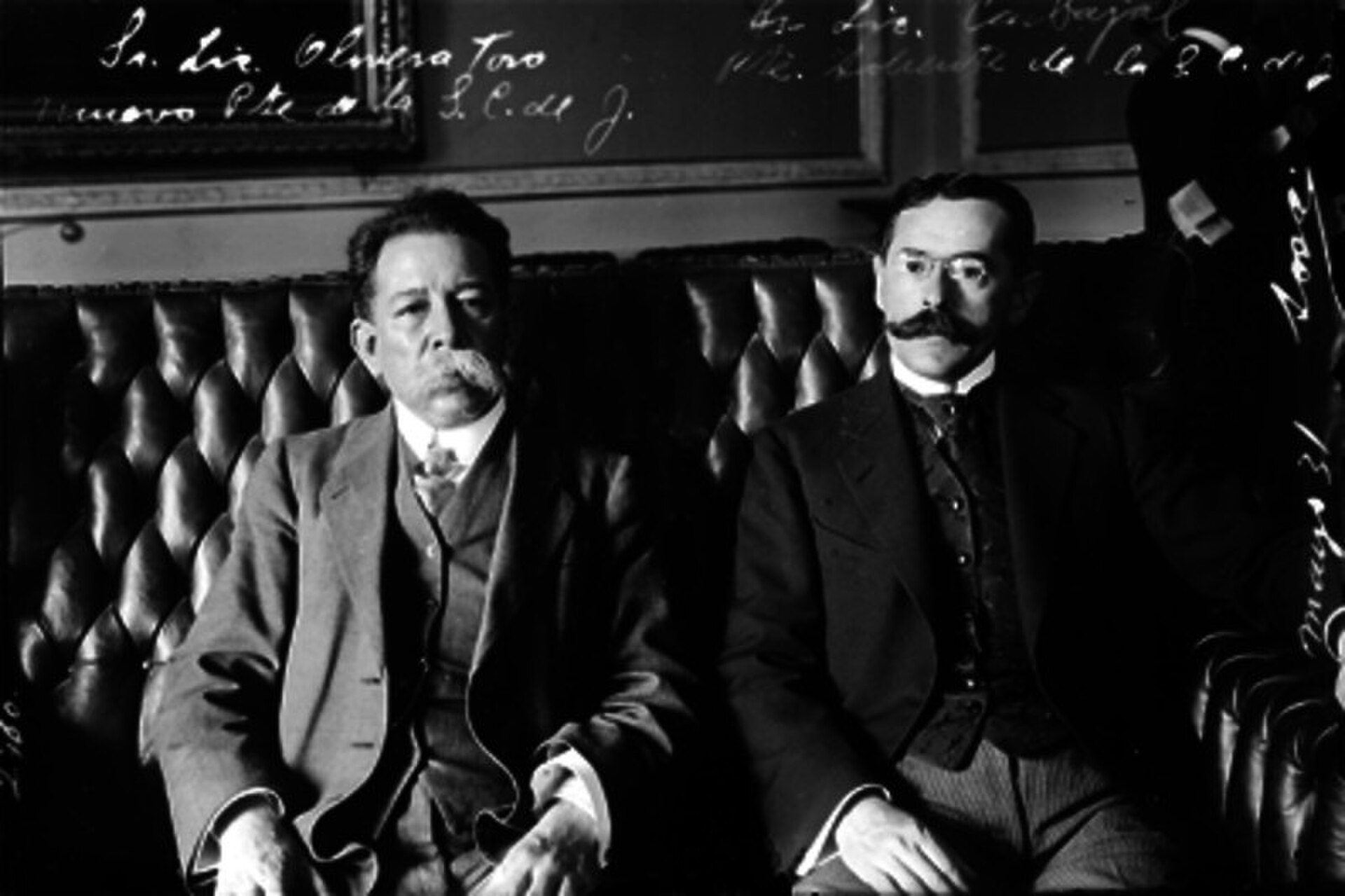 Two men in suits sitting on a leather couch in a vintage room.