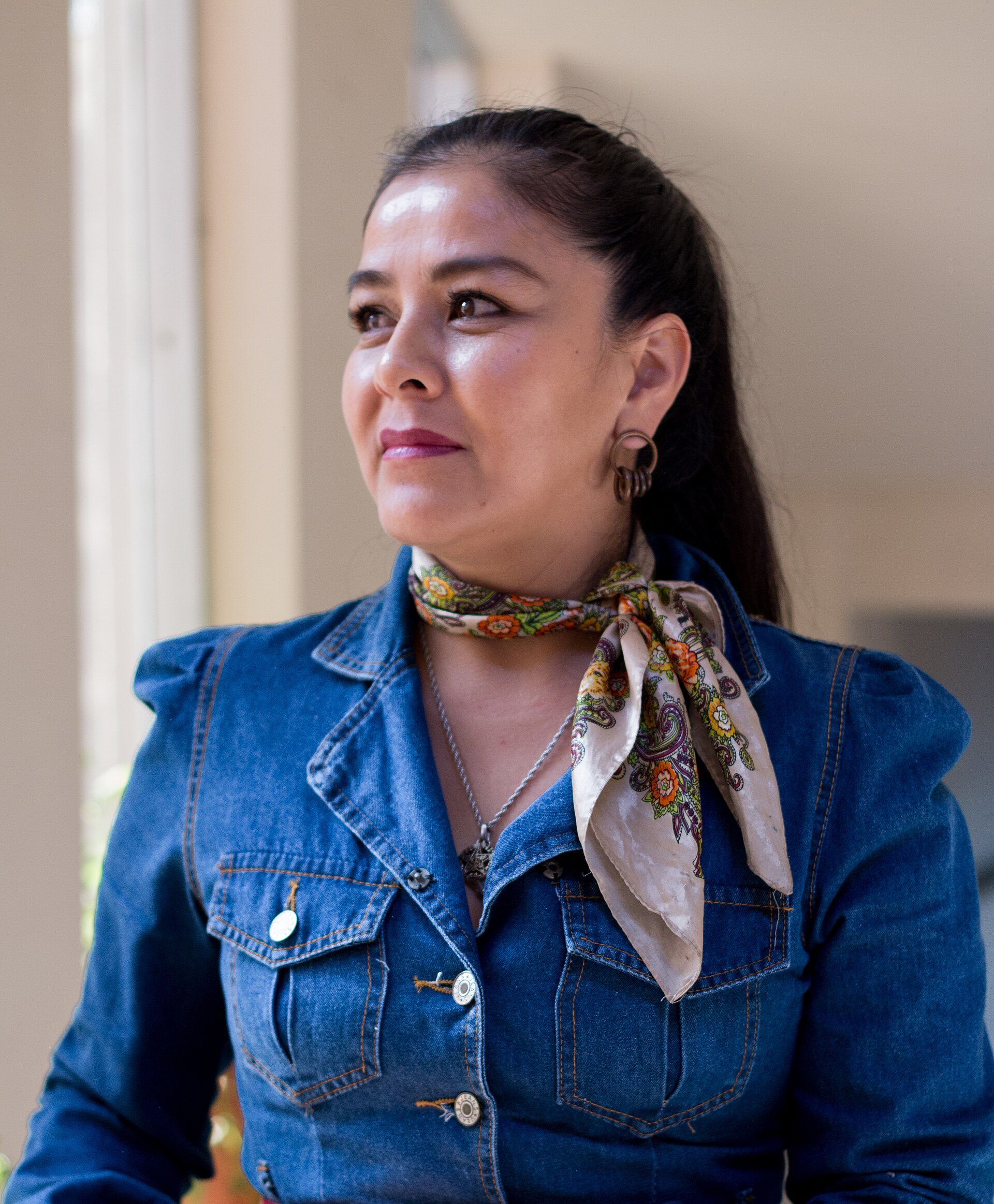 A woman in a denim jacket and scarf stands in a well-lit room with a modern interior.