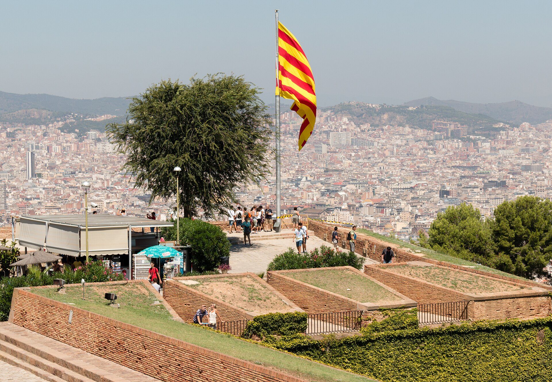 Panoramic city view from rooftop terrace with seating and a large flag.
