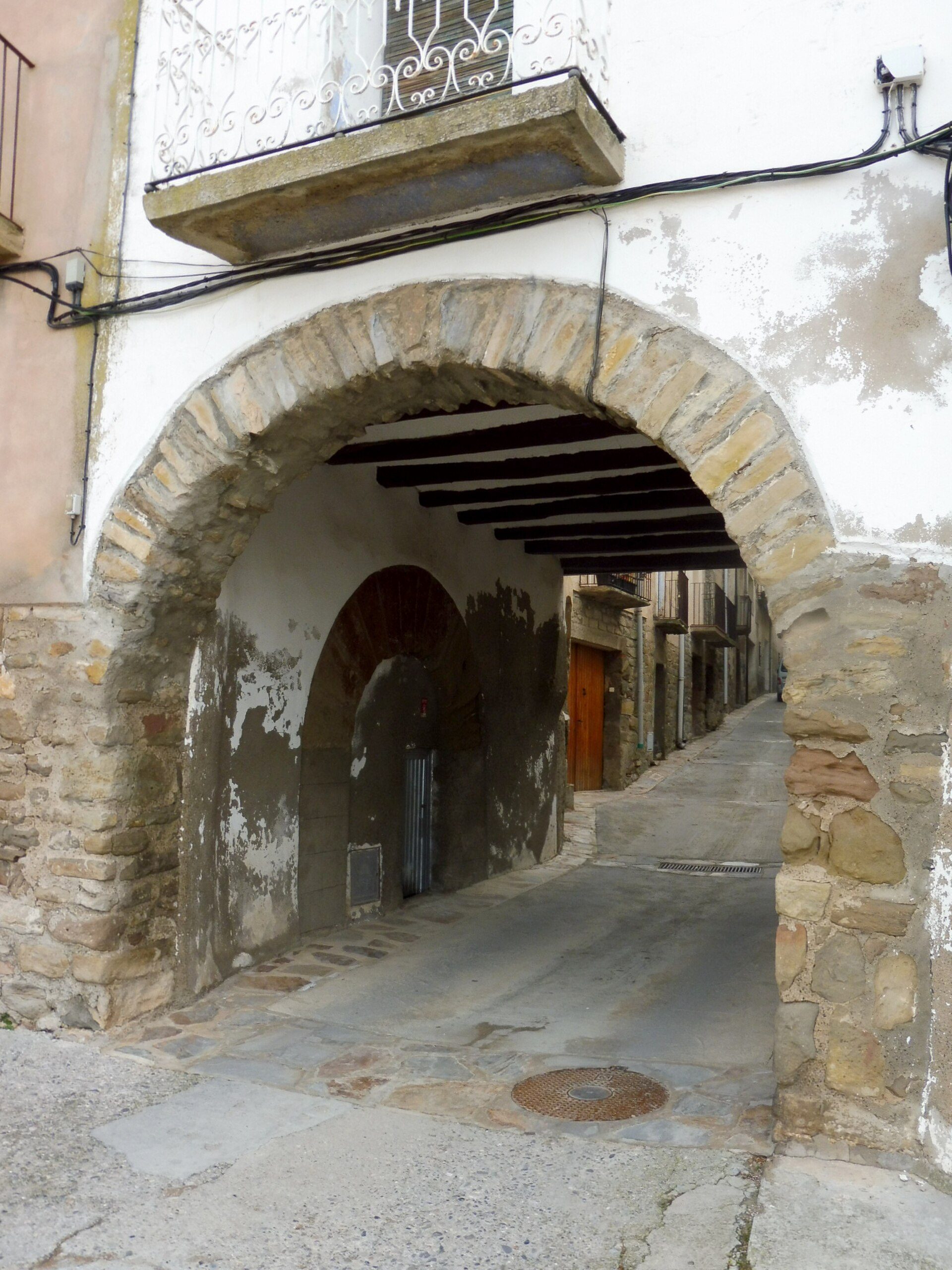 A stone archway leads to a narrow alley with a wooden door and a small balcony above.