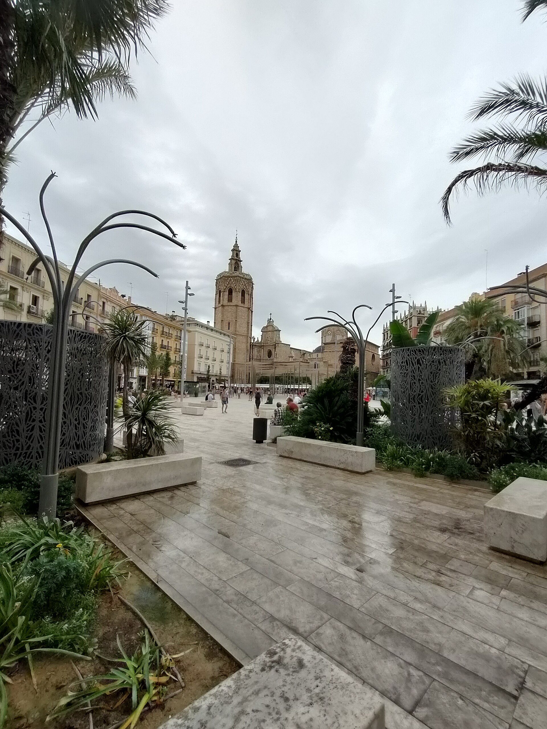 Alt text: Outdoor plaza with palm trees, decorative metal arches, and a historic church in the background.