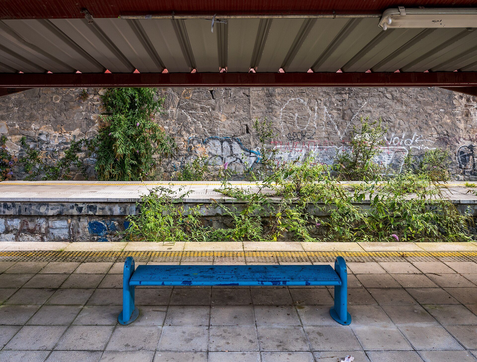 A blue bench on a train platform with a stone wall and greenery in the background.