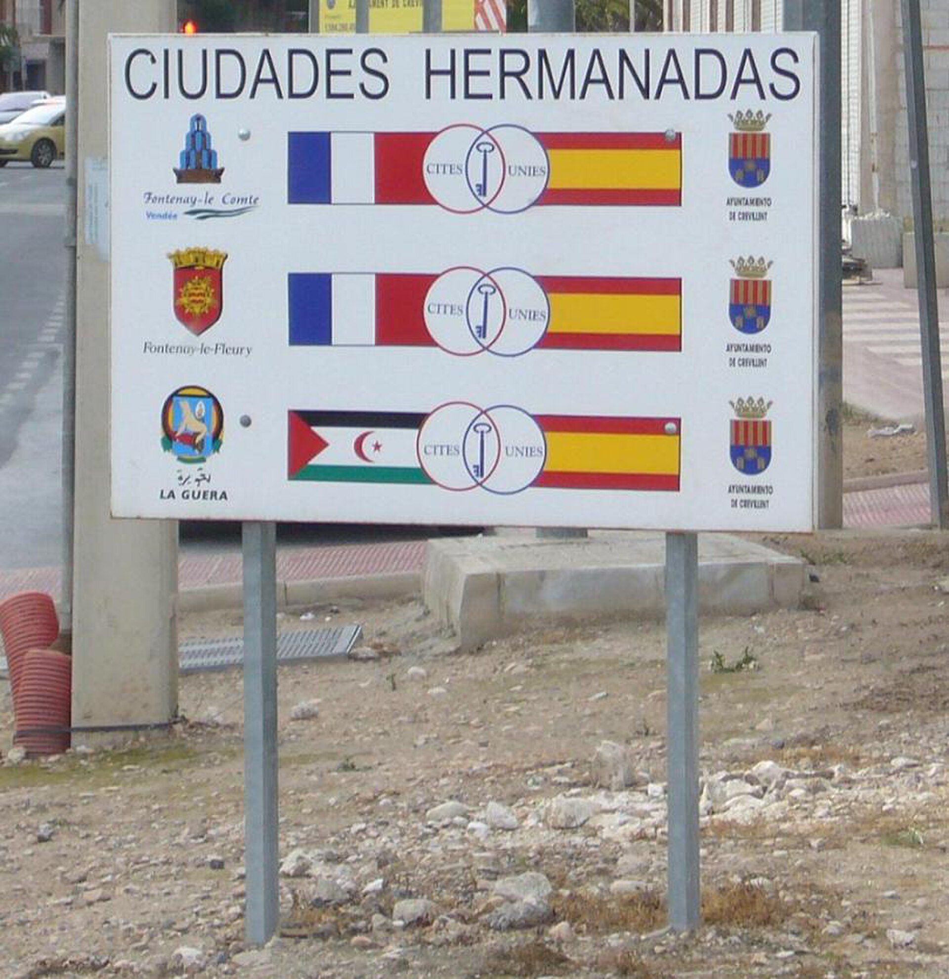 Signboard with city flags and emblems, located on a sidewalk.