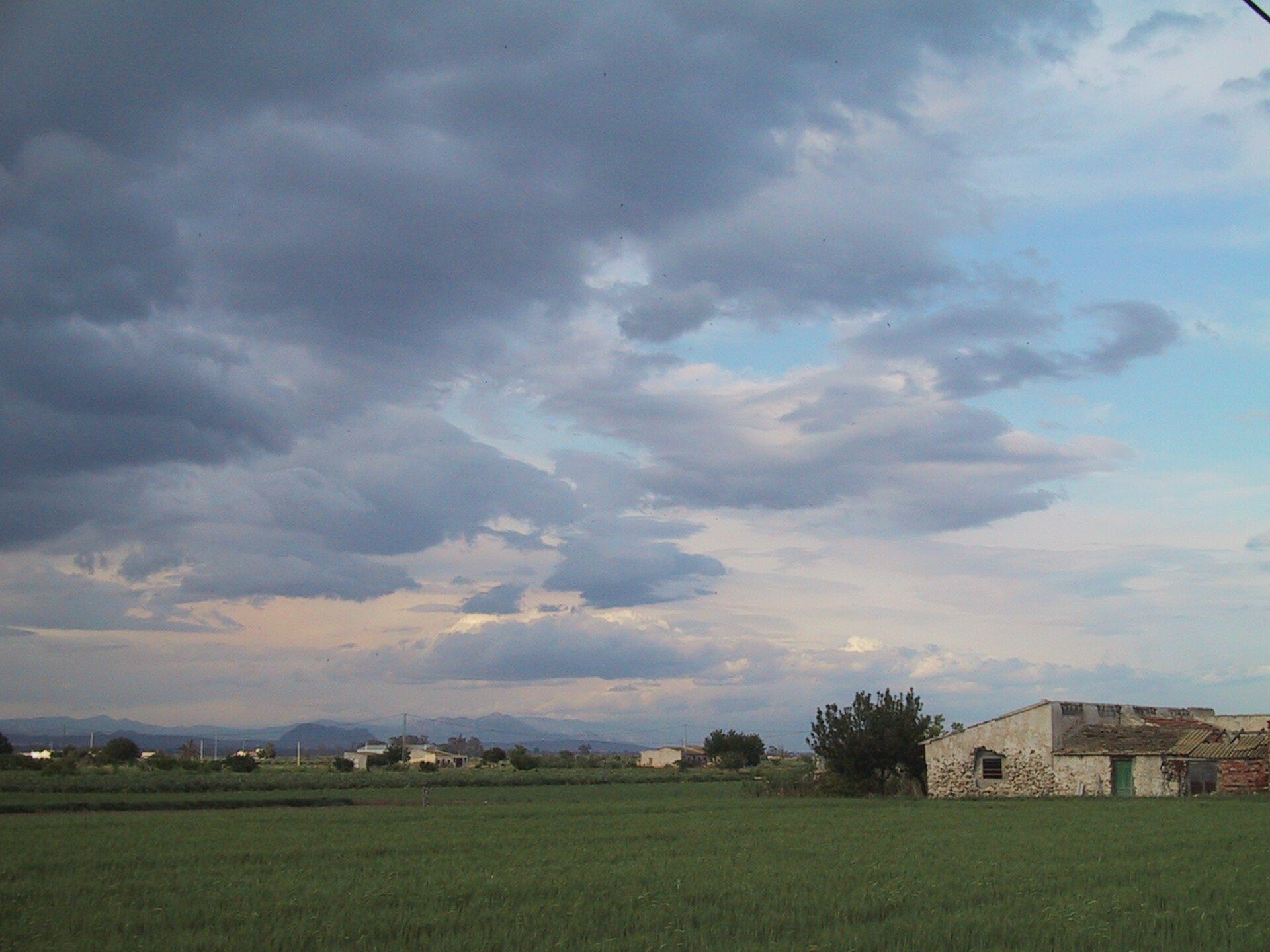 A rustic stone house with a field view, surrounded by greenery and distant mountains.
