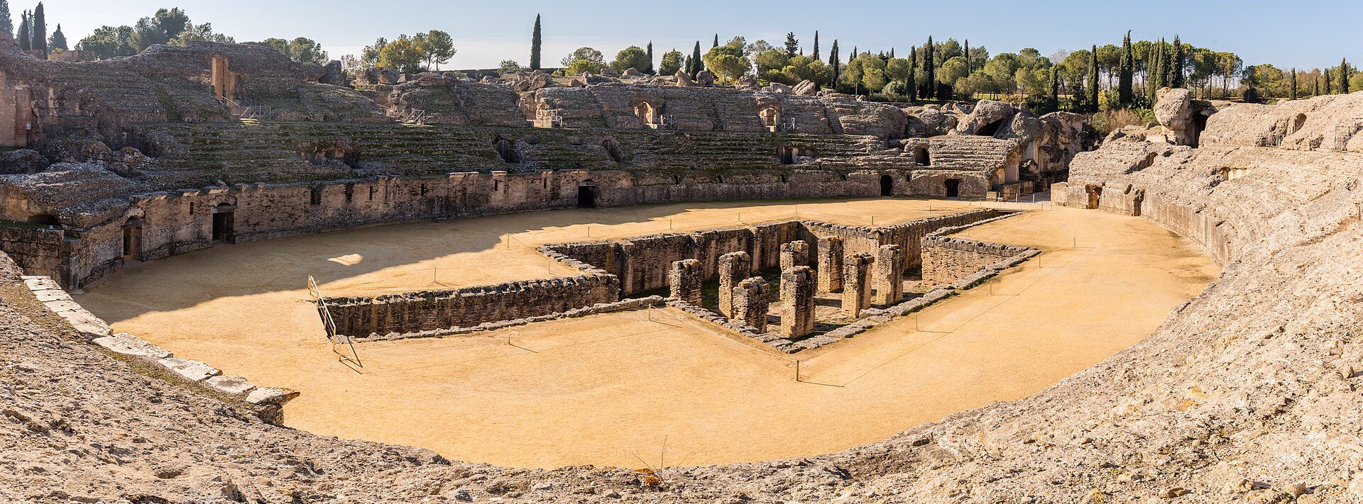 Ancient amphitheater with stone seating and central stage, surrounded by trees.