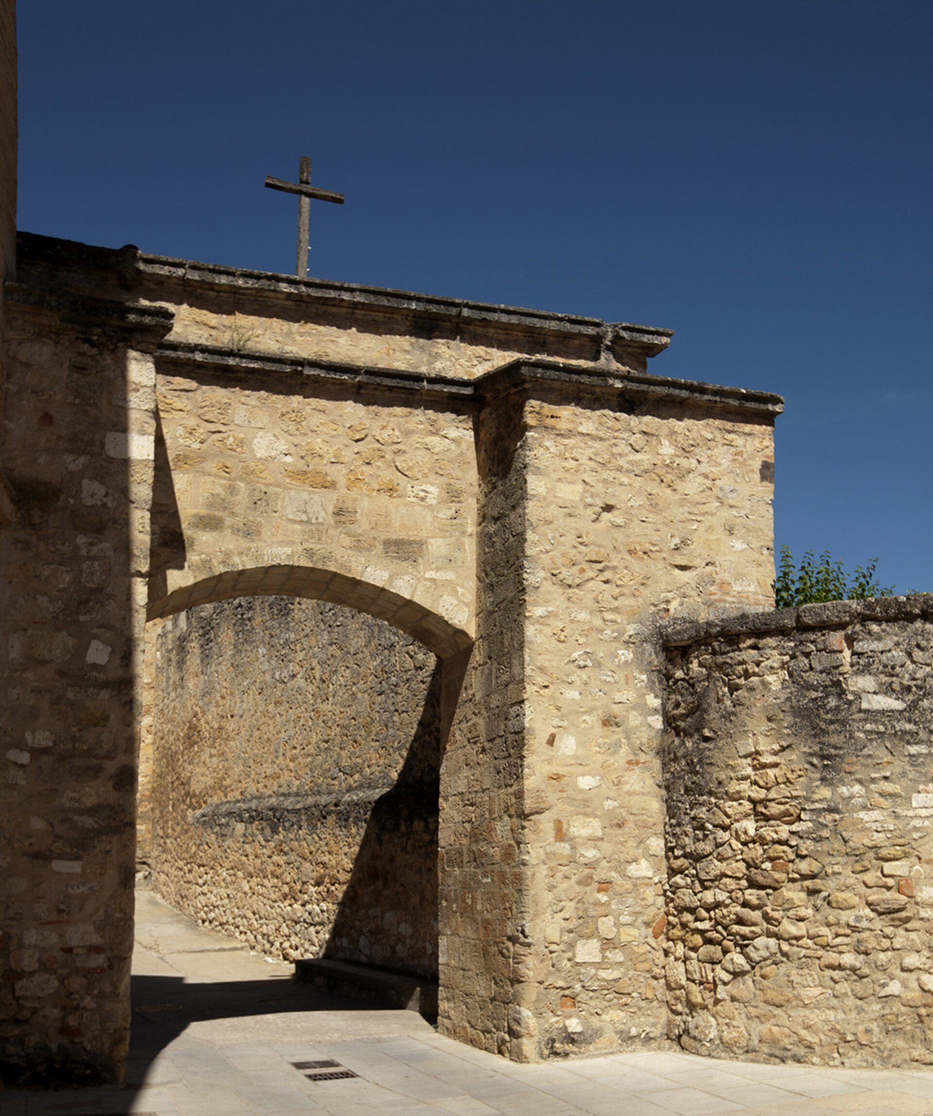 Stone archway with cross, leading to a courtyard, clear blue sky.