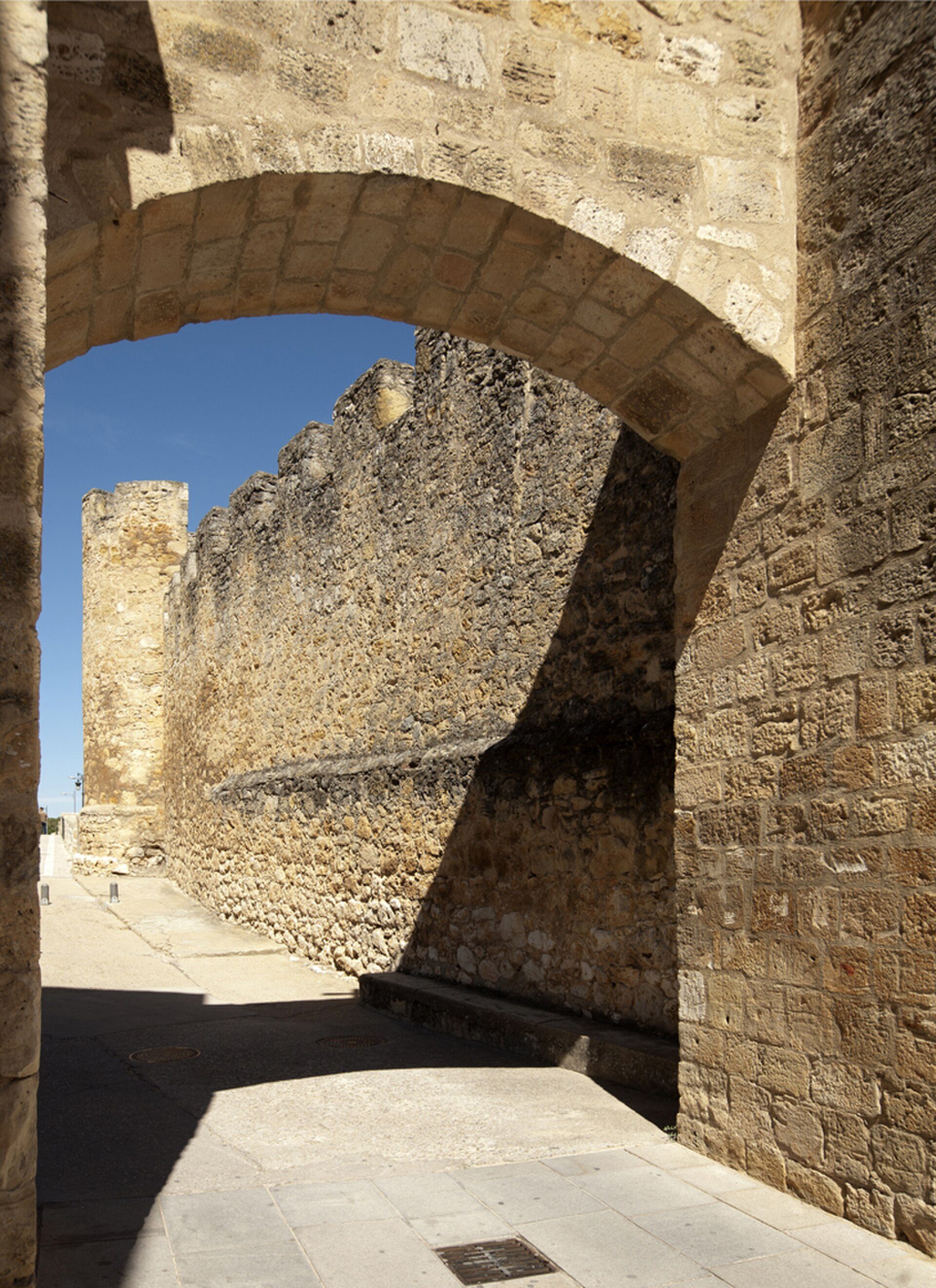 Stone archway leading to a historic stone wall and tower, with a clear blue sky view.
