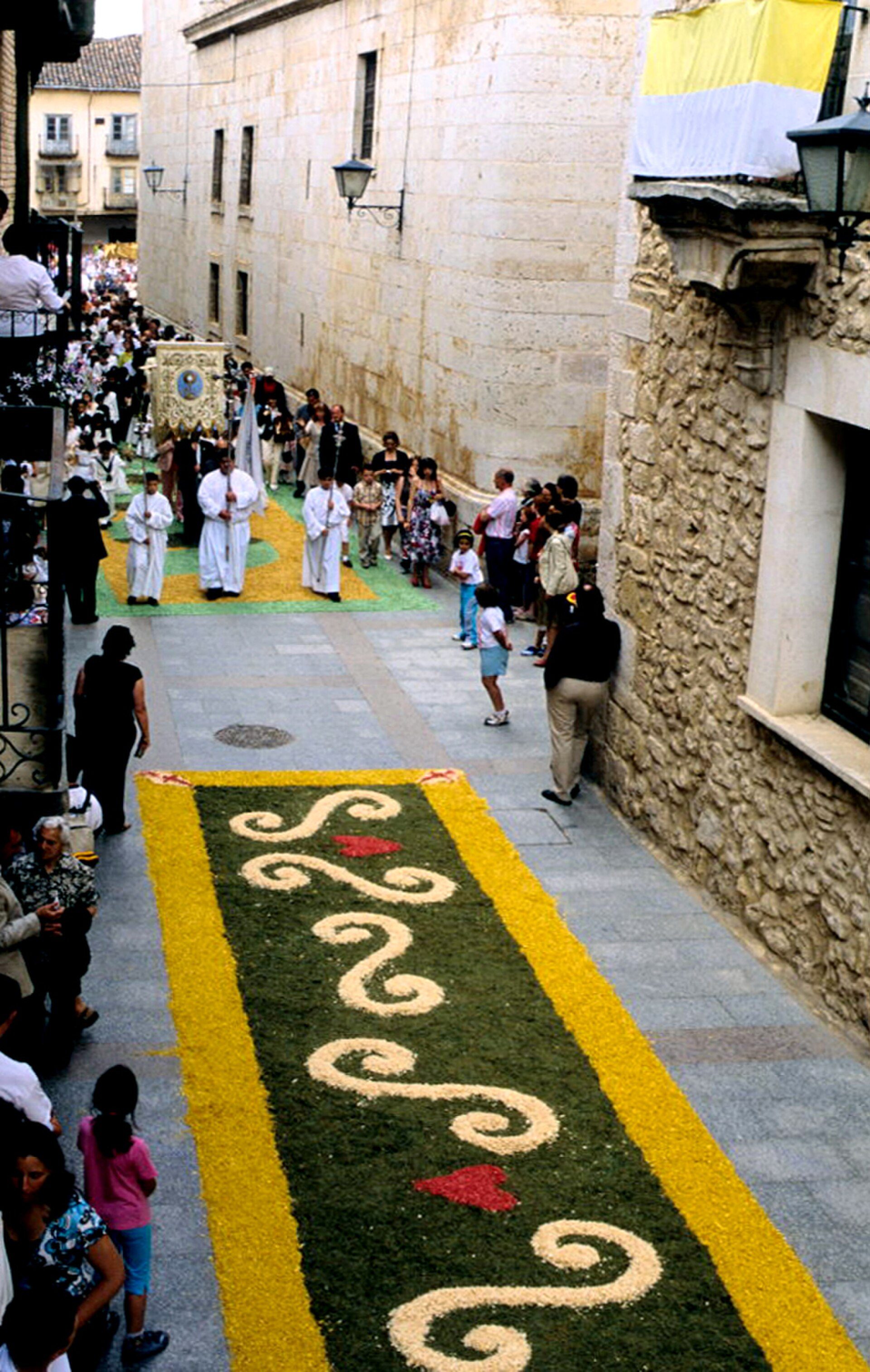 Alt text: Narrow street with a floral carpet, people in procession, stone buildings.