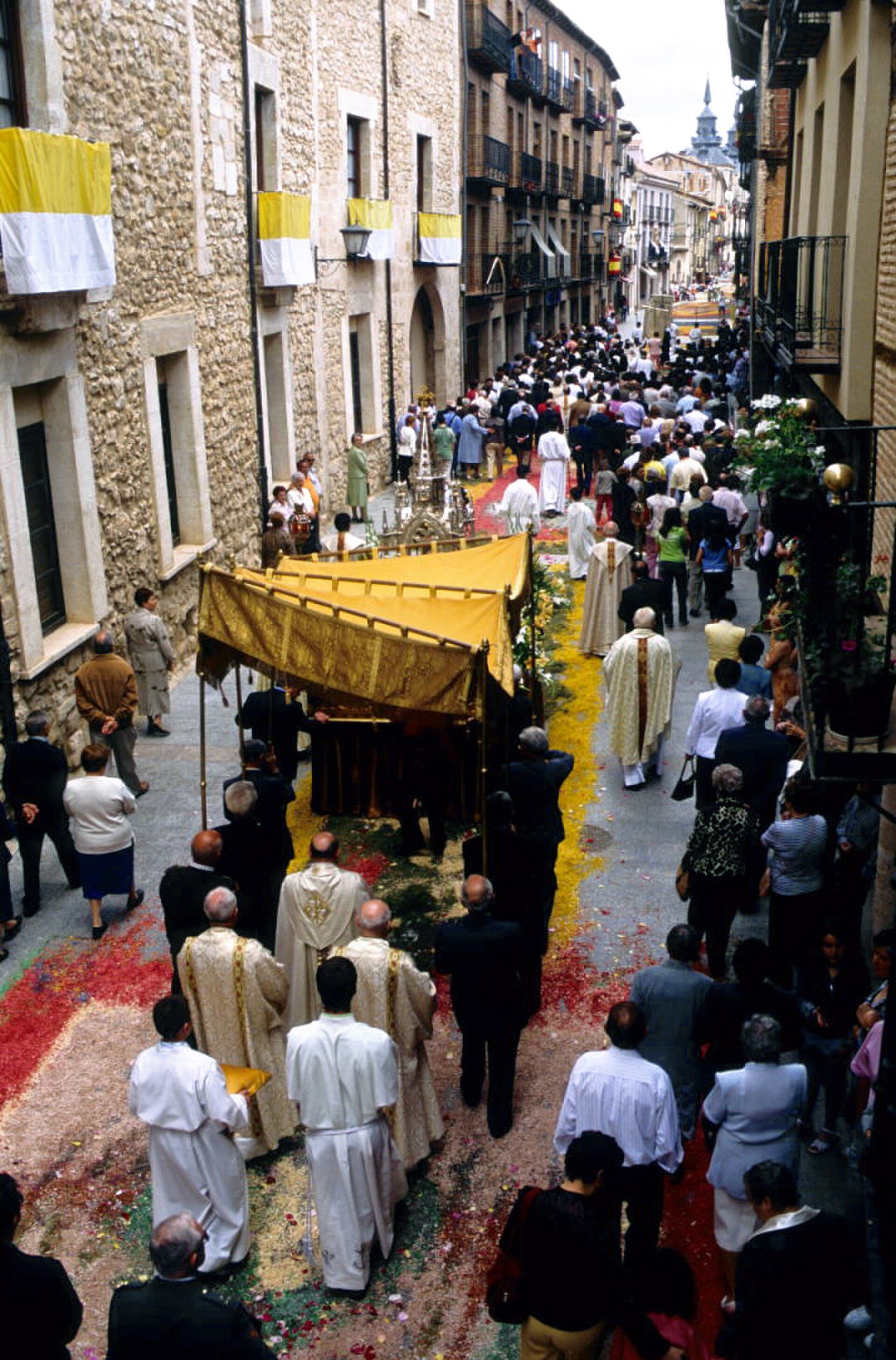Alt text: Street procession with clergy in white robes, colorful confetti, and onlookers.
