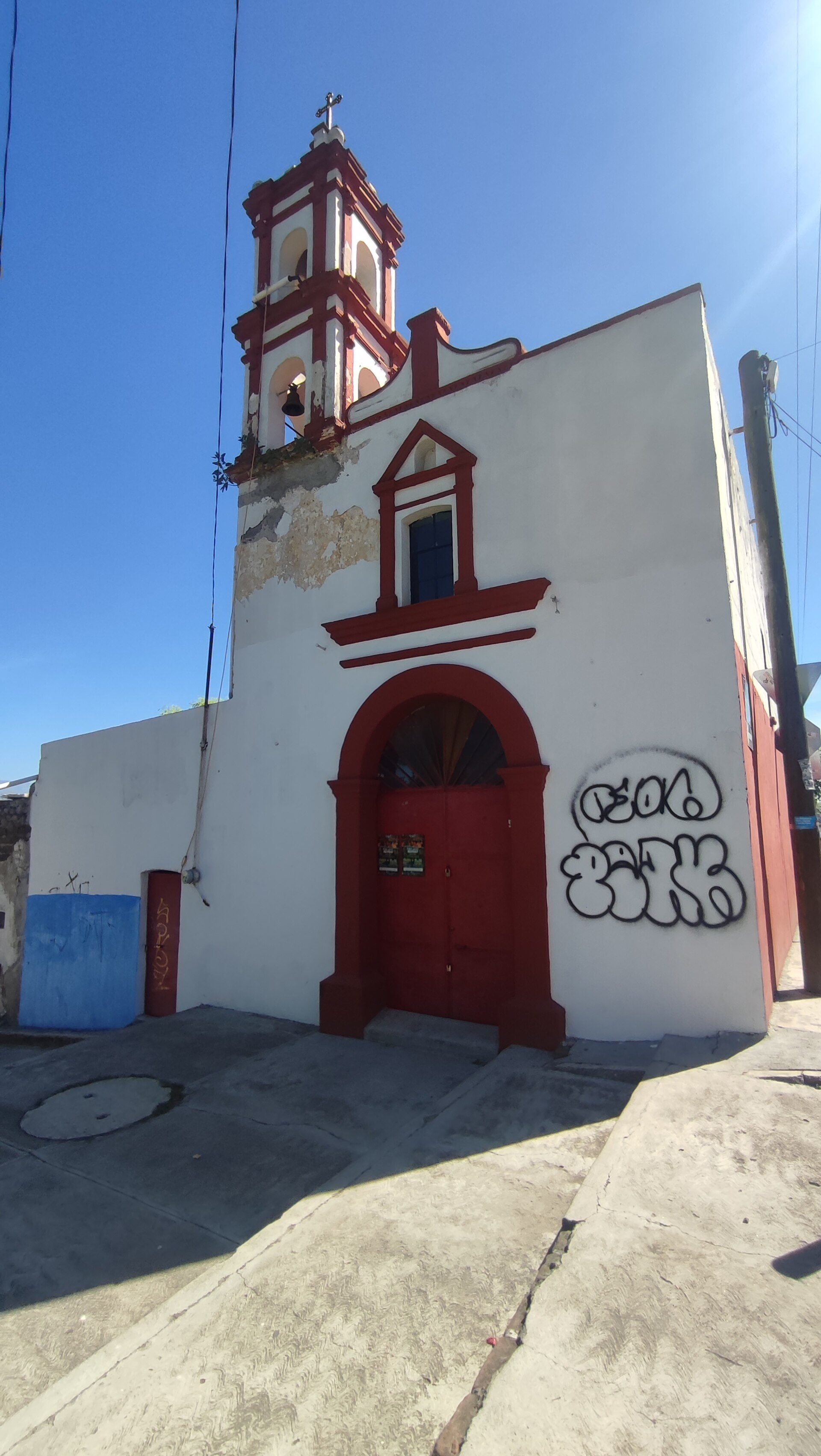 Small church with red doors, white walls, and a bell tower, set against a clear blue sky.