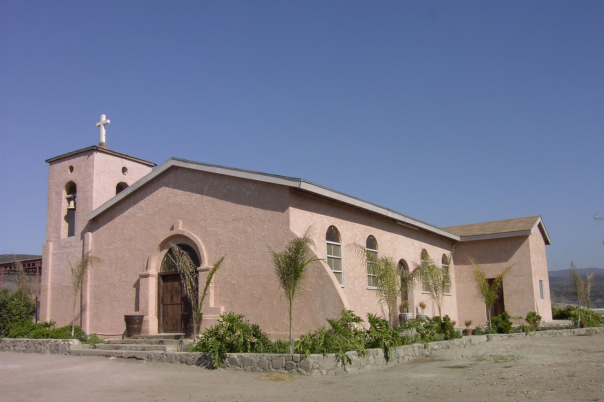 A small church with a cross on top, surrounded by greenery and a clear blue sky.