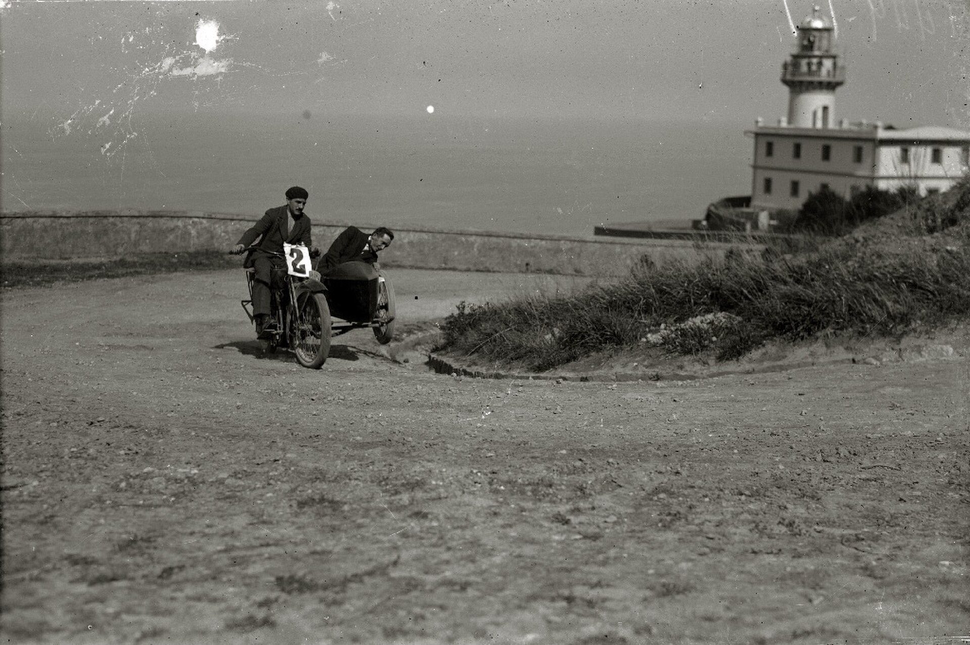 A vintage black and white photo of a motorcycle race on a coastal road with a lighthouse in the background.