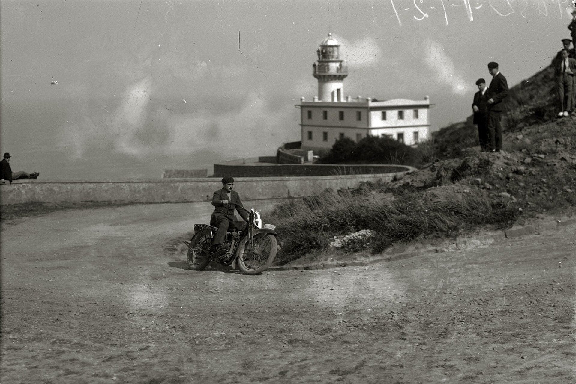 A vintage black-and-white photo of a man riding a motorcycle near a lighthouse with spectators.