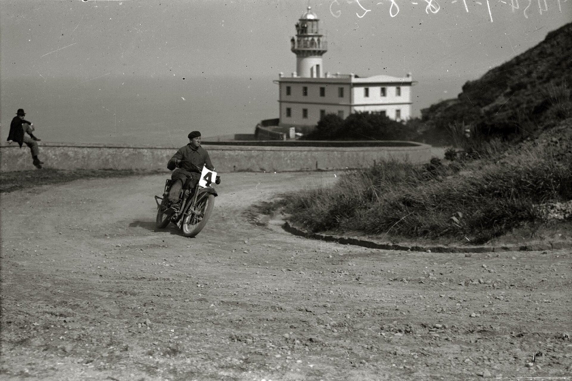 A vintage black-and-white photo of a motorcycle on a coastal road with a lighthouse in the background.