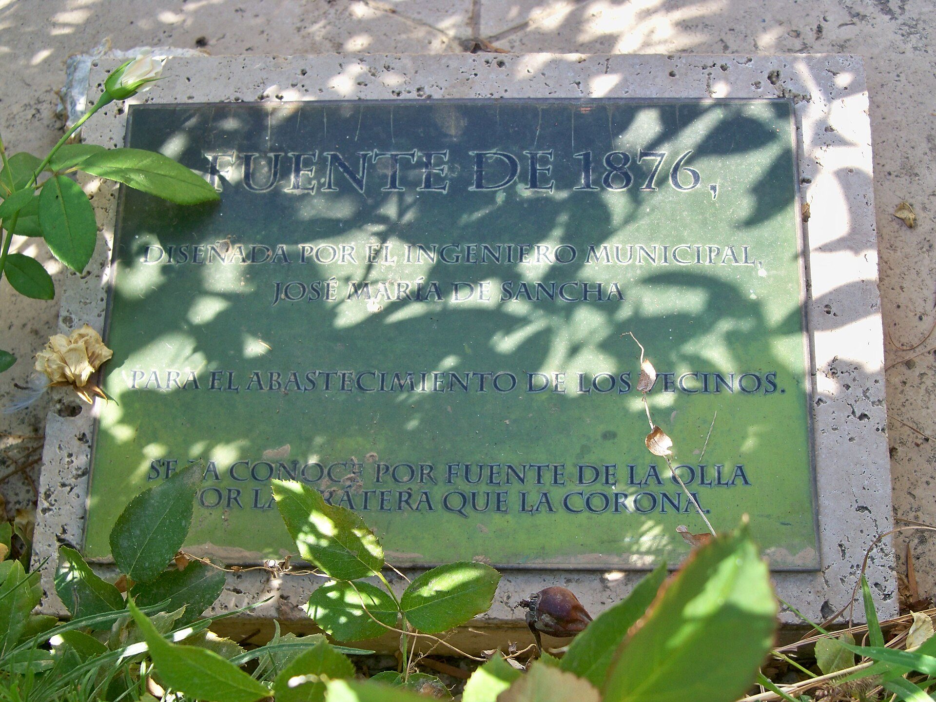Alt text: Green plaque commemorating the 1876 Fuentes de Alaola water fountain, surrounded by plants.