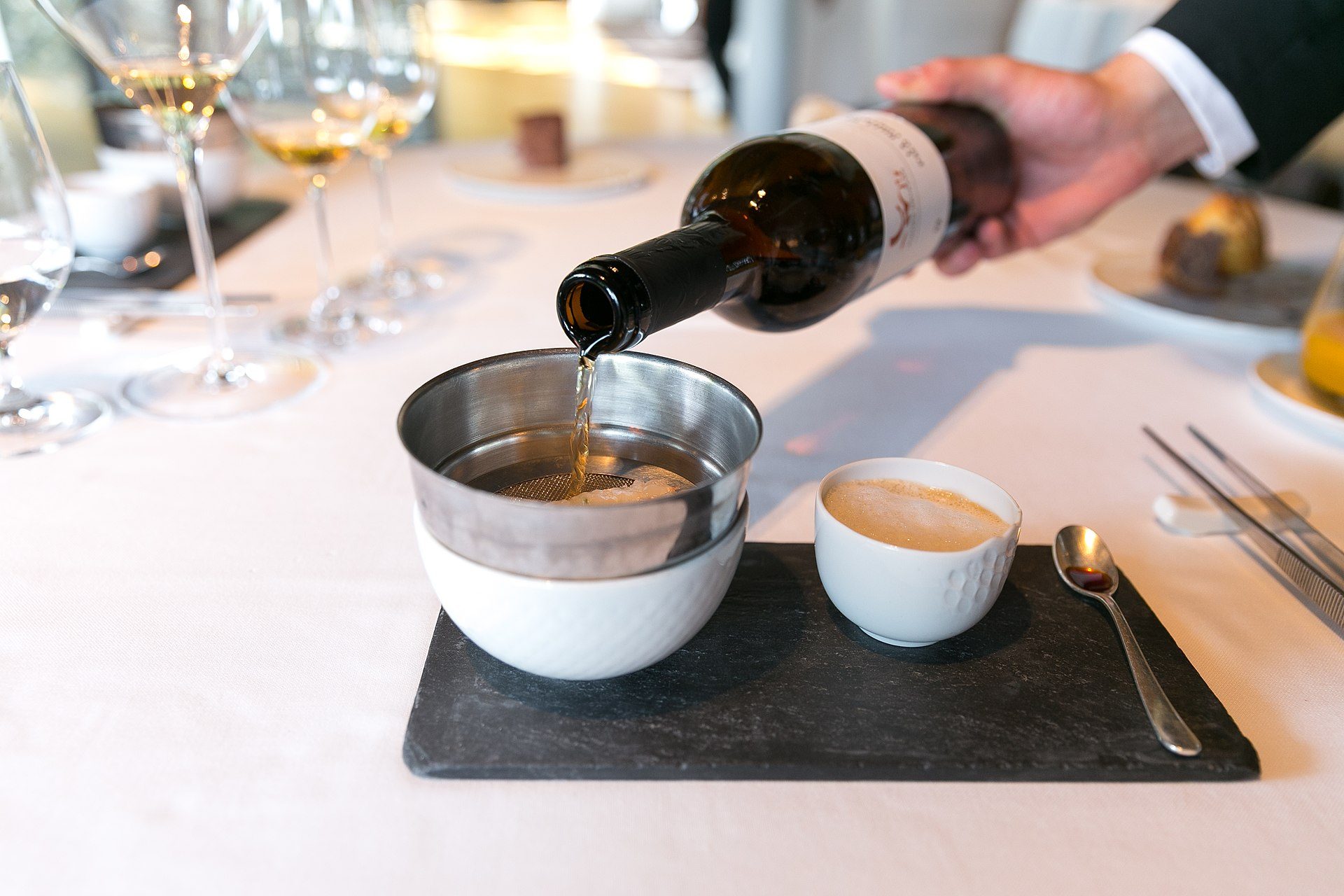 A hand pouring wine into a bowl on a dining table with glasses and cutlery.