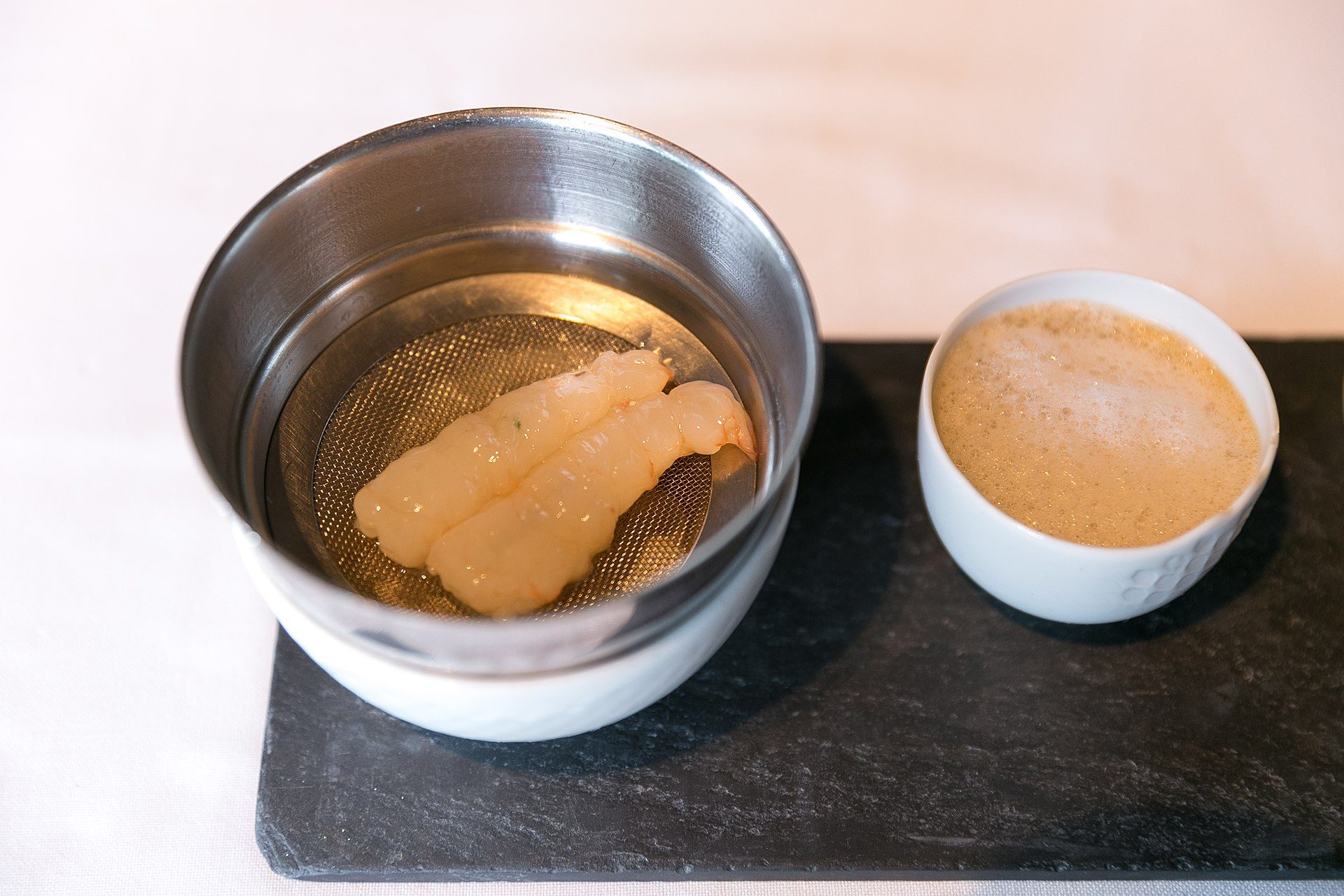 Steaming seafood in a metal bowl with a small cup of sauce on a black tray.