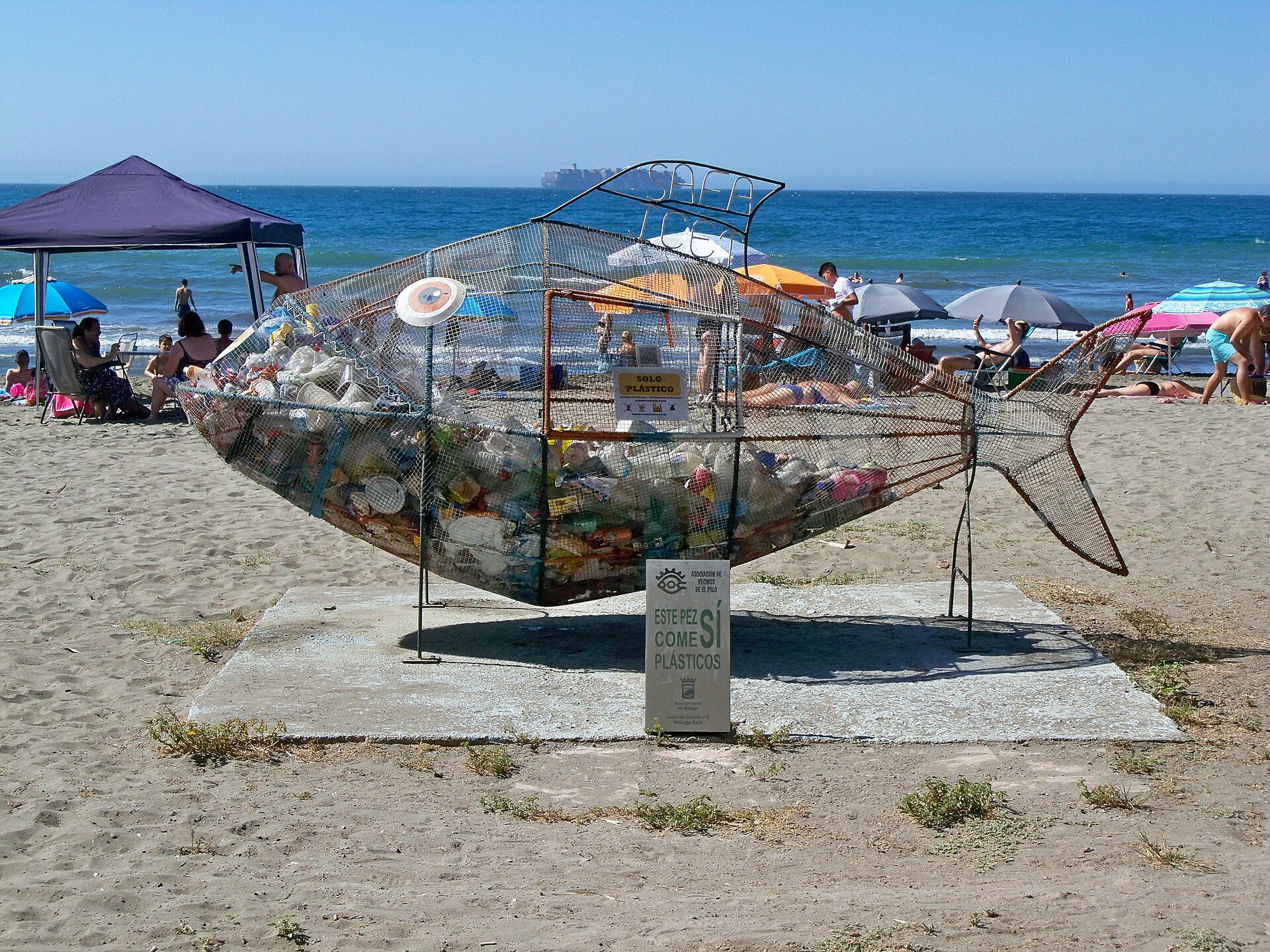 Beach sculpture with fish design, filled with plastic waste, on sandy shore with ocean view.