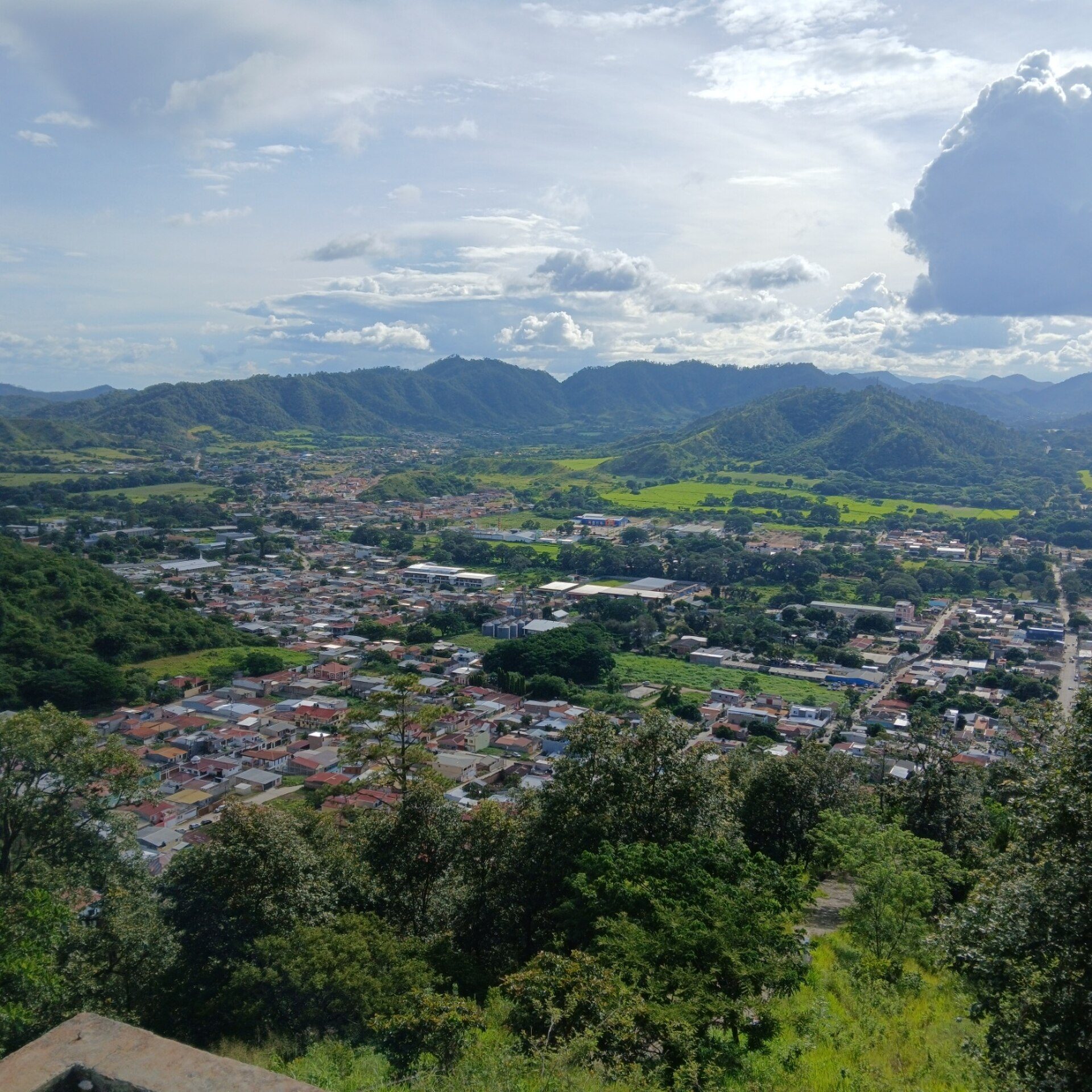 Alt text: Panoramic view of a small town with lush green hills and scattered clouds.