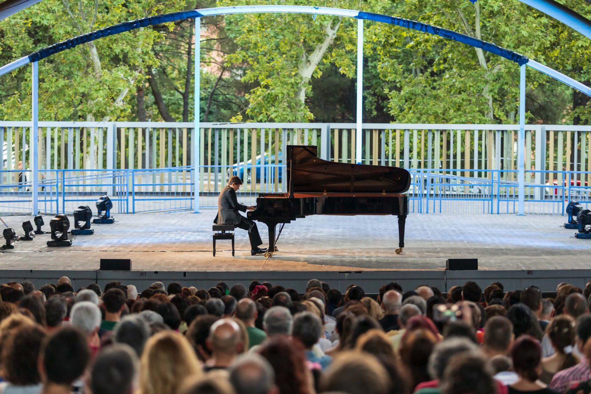An outdoor concert scene with a pianist on stage, surrounded by a large audience.