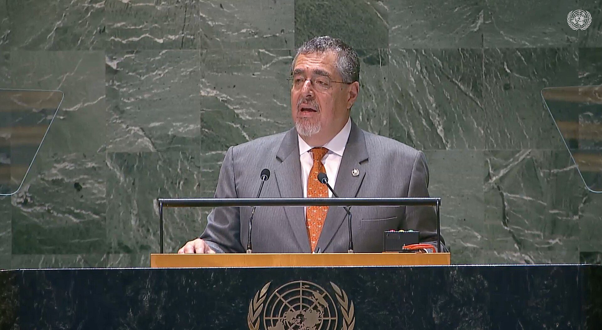A man in a suit and tie speaks at a podium in a formal conference room with a marble background.