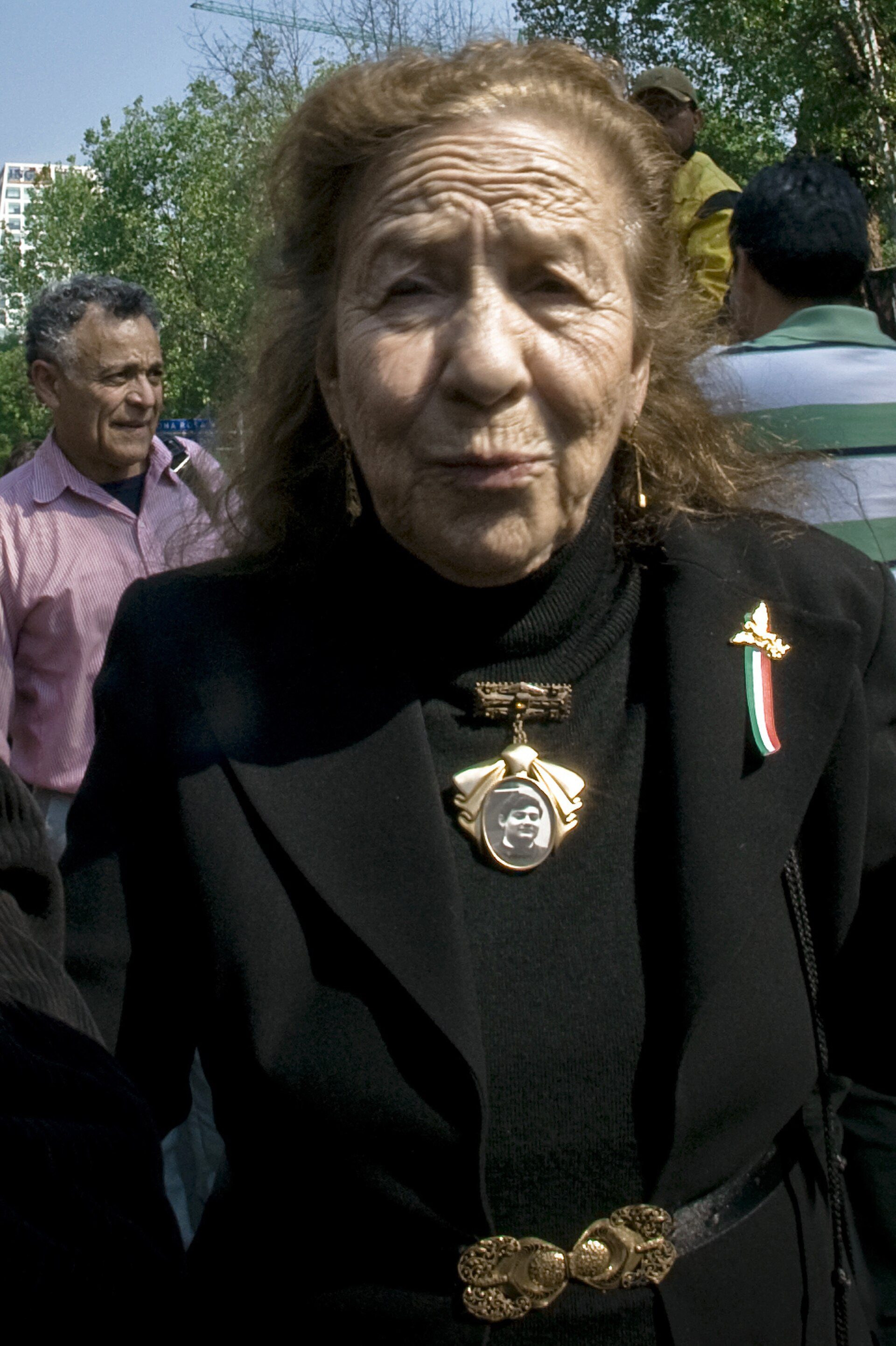 Alt text: Elderly woman in black blazer with medals, standing outdoors.