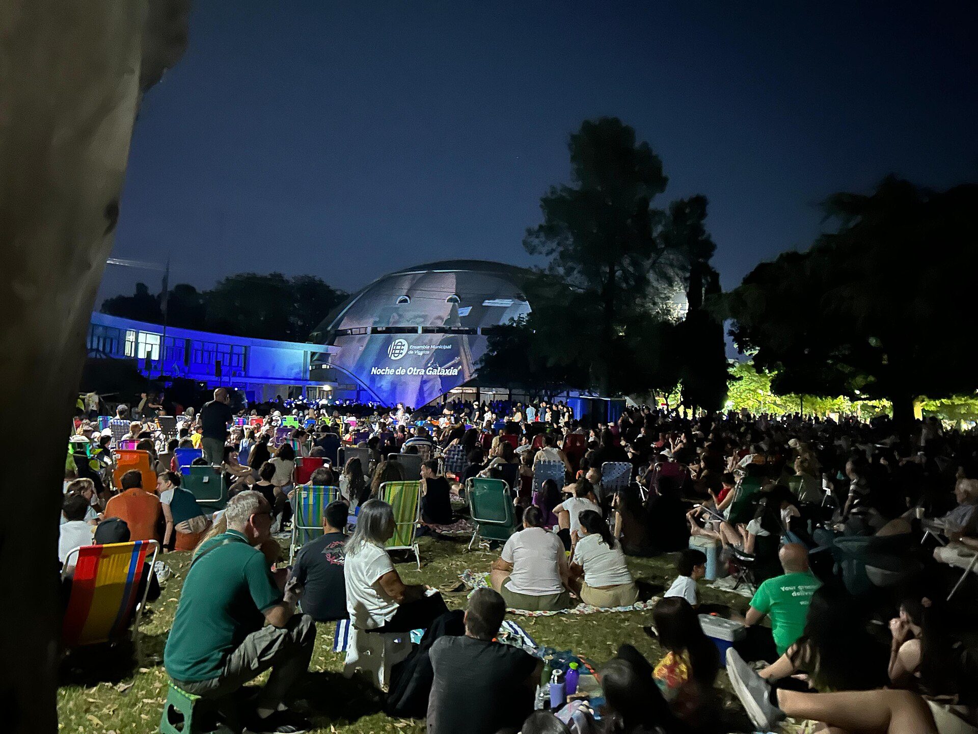 Alt text: Outdoor concert at night with a large crowd seated on chairs and blankets, facing a stage with a dome structure in the background.