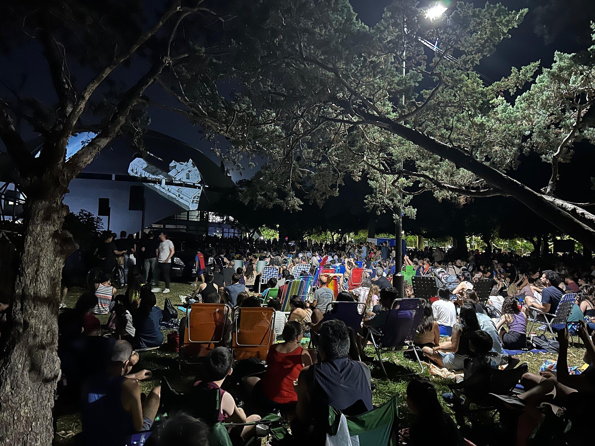Outdoor movie night with a large crowd, trees, and a large screen.